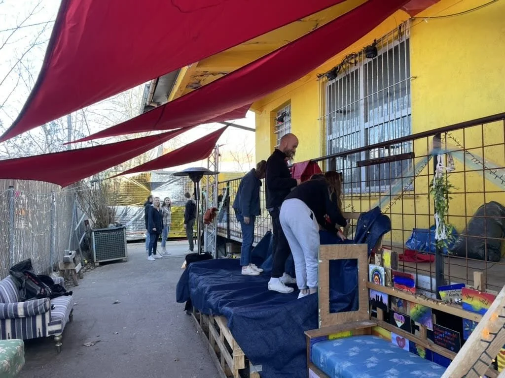 People shopping at an outdoor market with a yellow building, red canopy, and various items for sale, including paintings and artwork.