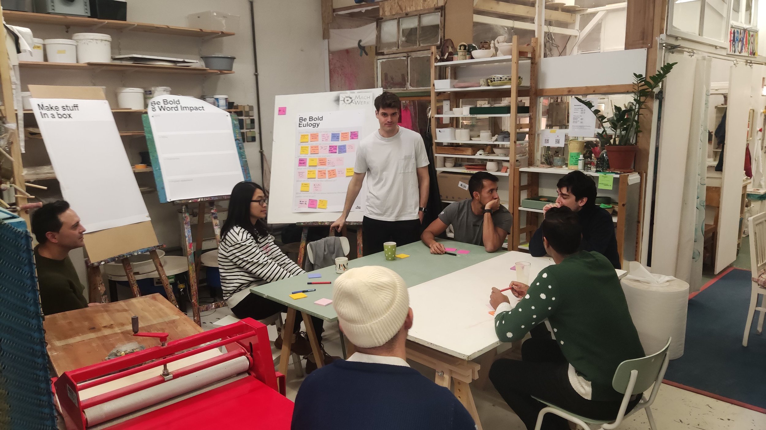 Group of people participating in a workshop or meeting in a cozy, industrial-style room with shelves, plants, and boards with colorful sticky notes and writing.