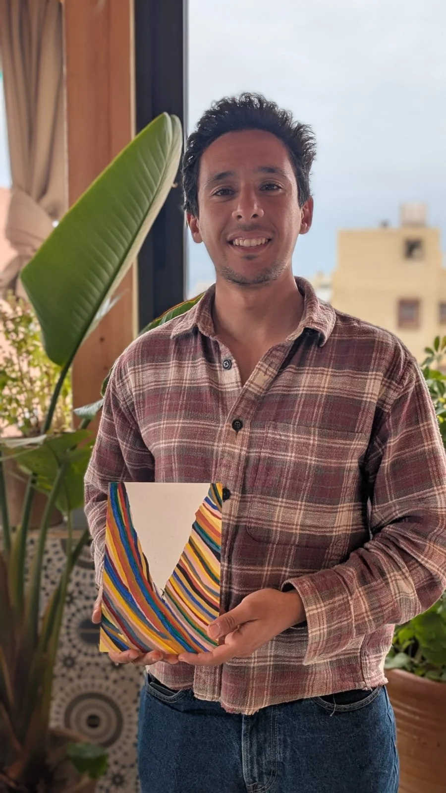 A smiling man with dark curly hair and light facial hair holding a colorful abstract painting, standing indoors near large windows with plants and city buildings in the background.