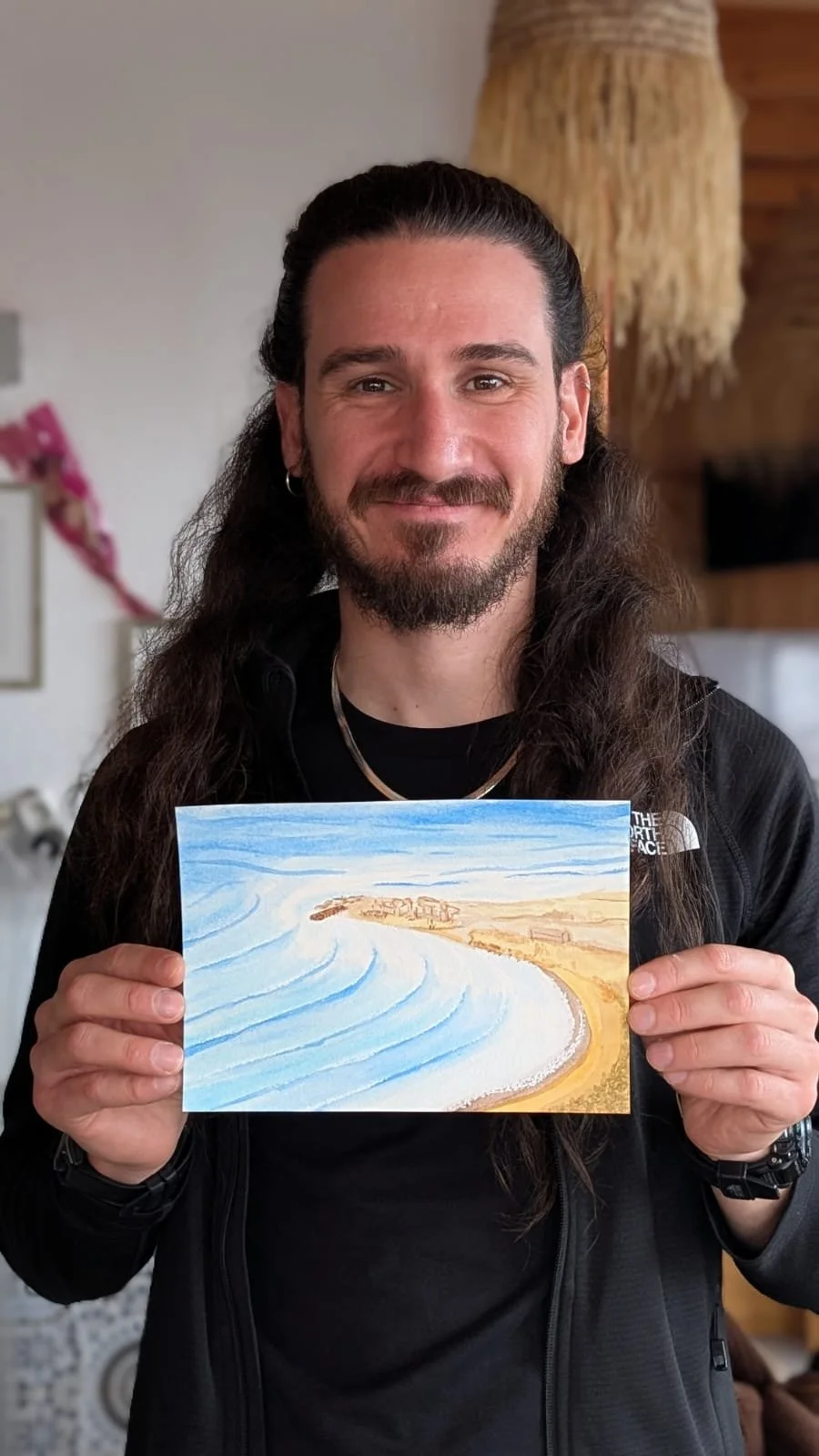 A man with long dark hair, beard, and mustache, smiling, holding a painting of a coastal landscape with waves and beach.