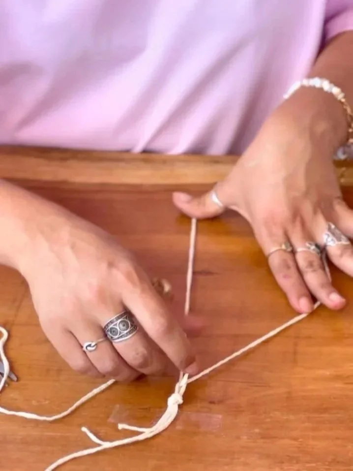 Person's hands tying a knot in a piece of string on a wooden surface, wearing multiple rings and a bracelet.