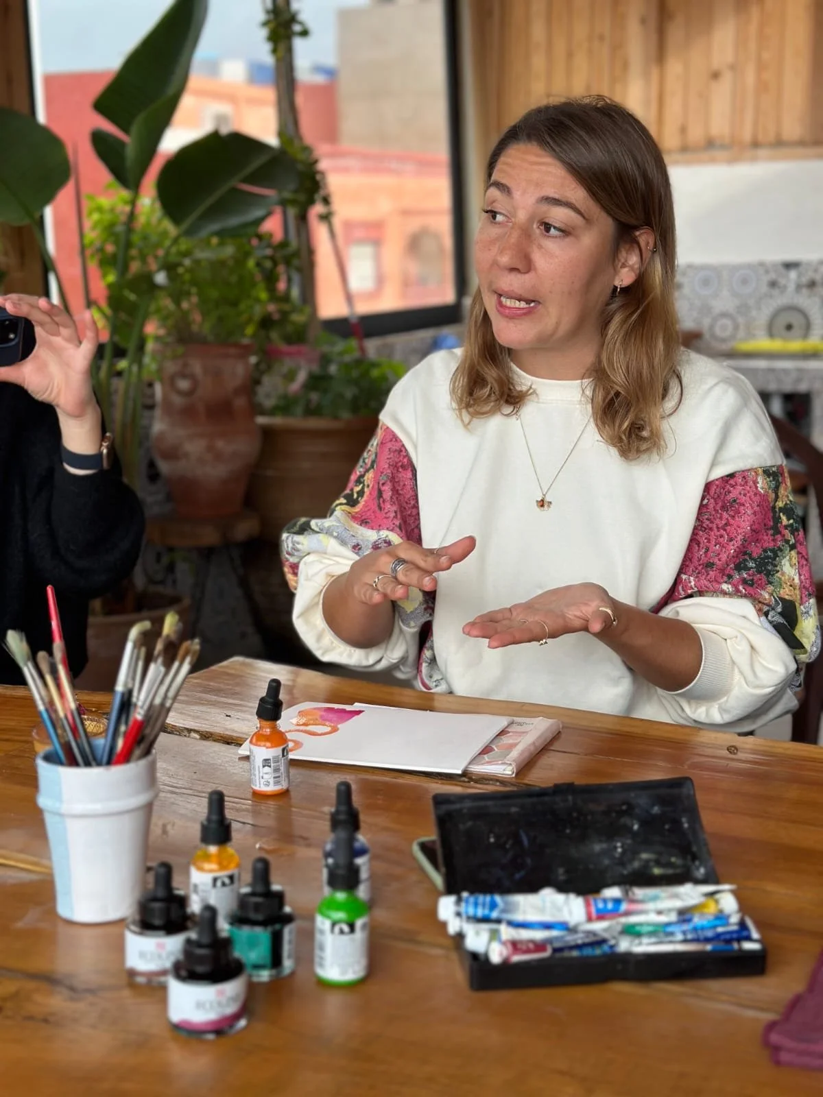 A woman sitting at a wooden table, gesturing with her hands, surrounded by art supplies including bottles of paint, brushes in a container, and tubes of paint.
