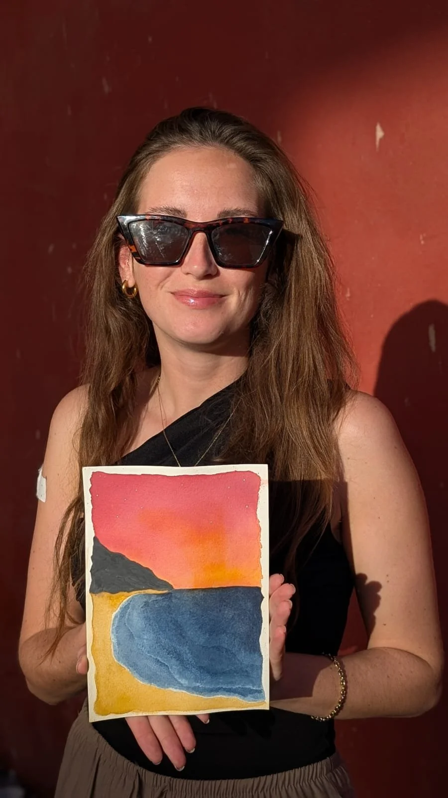 A woman with long brown hair, wearing sunglasses, gold hoop earrings, and a black top, holding a watercolor painting of a beach and sunset scene, standing against a red wall.