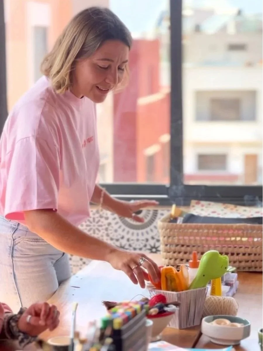 A woman wearing a pink t-shirt and light jeans smiling and organizing arts and crafts supplies on a table near a window.