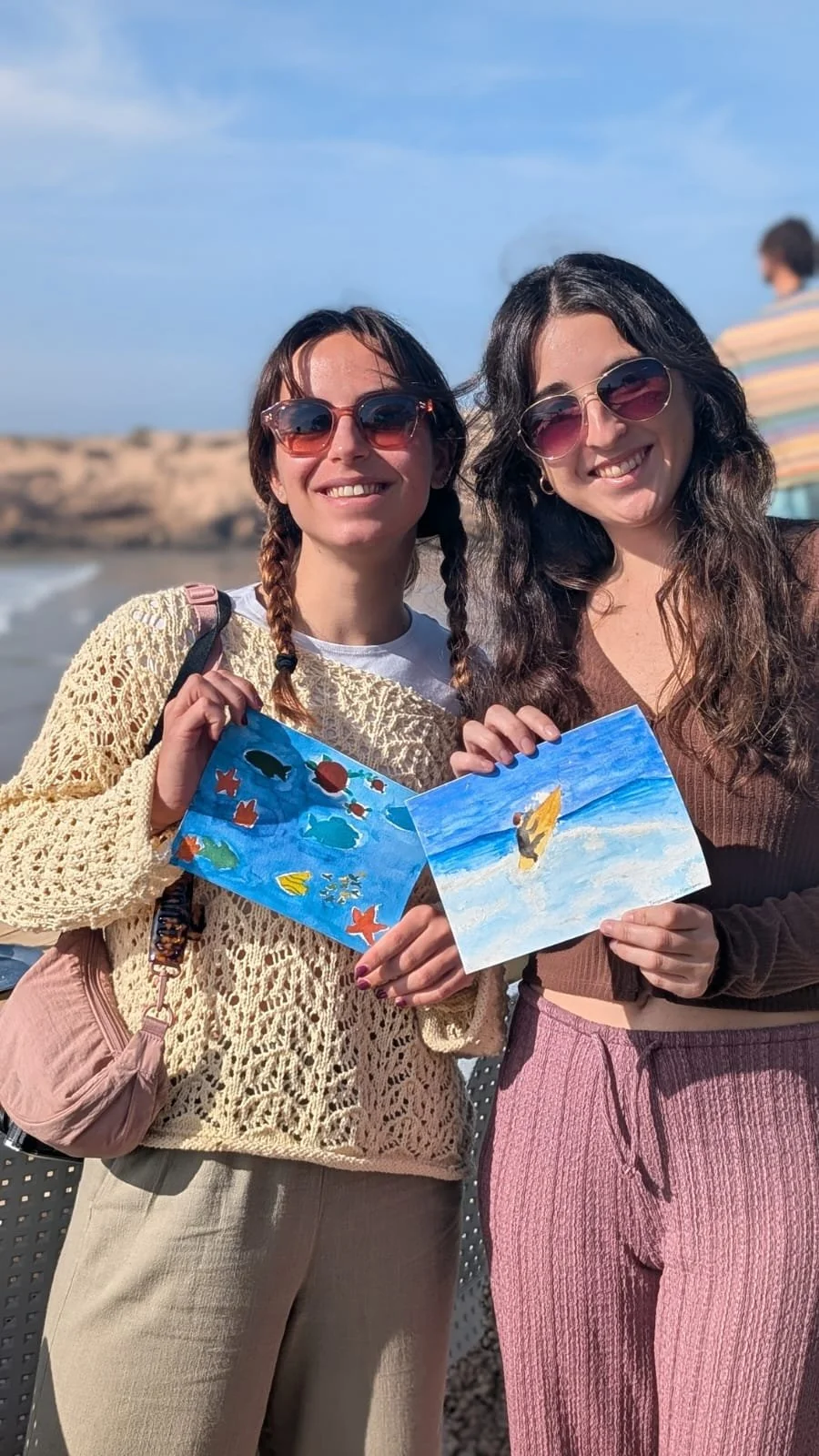 Two women wearing sunglasses holding paintings of an ocean scene on a beach, smiling at the camera with a rocky shoreline and blue sky in the background.