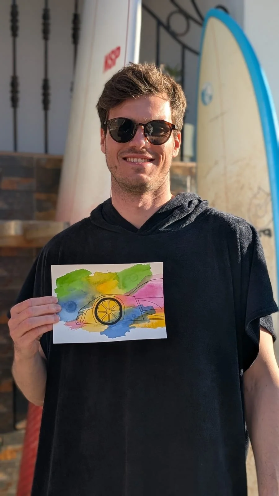 A man wearing sunglasses, smiling, wearing a black hoodie, holding a colorful watercolor painting of a car with a lemon slice on its front, with a background of green, yellow, pink, and blue colors.