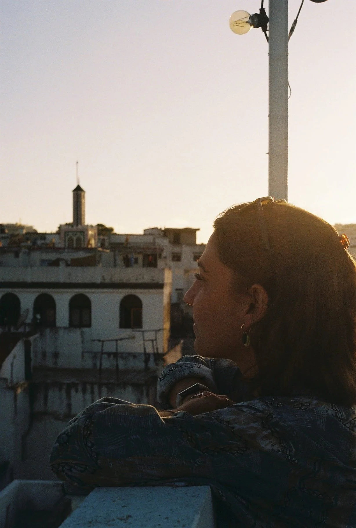 A woman standing on a rooftop at sunset, looking to the side with her arms crossed, with a cityscape and a streetlight in the background.