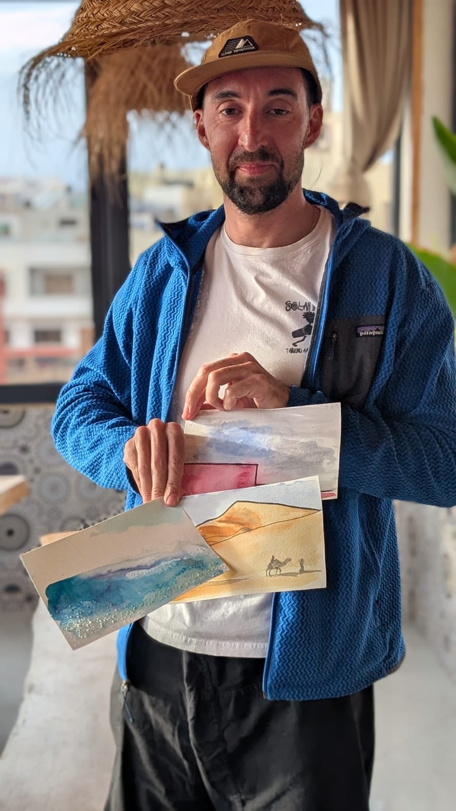 A man in a blue jacket and white shirt holding several watercolor paintings, standing inside a room with a balcony view of buildings outside.