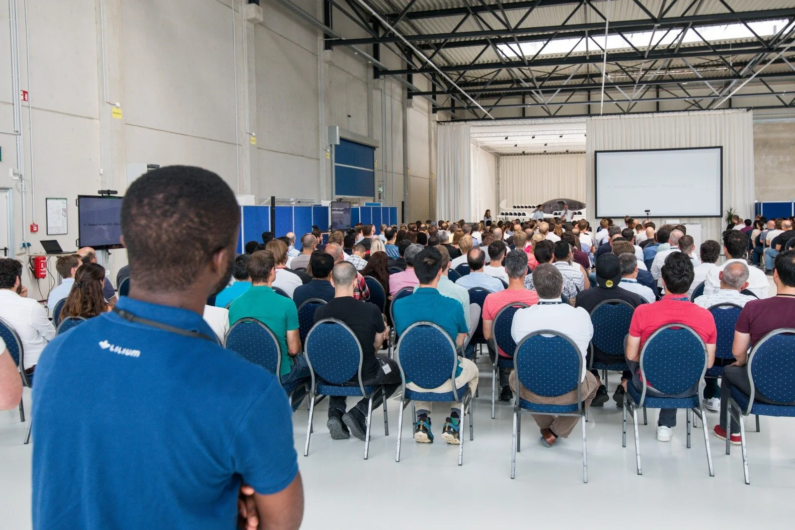 Large audience attending a conference or presentation in an industrial-style venue, with a speaker and a large screen at the front.