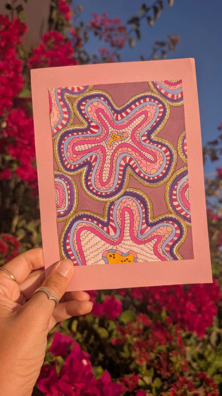 Hand holding a pink-bordered card with an abstract, colorful, and dotted pattern resembling Rorschach inkblot images against a background of pink flowers and a clear blue sky.
