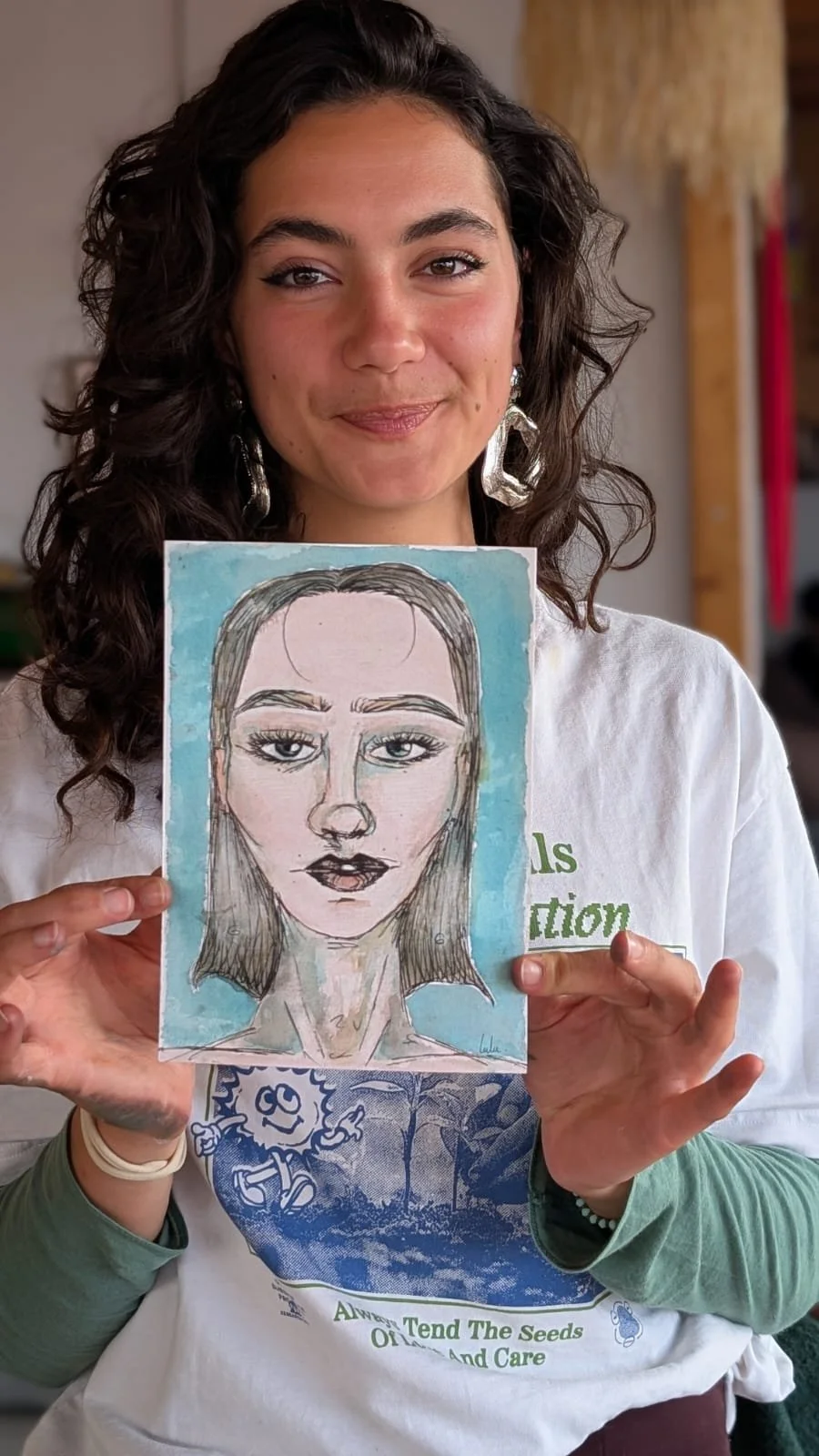 A woman with dark, curly hair is holding a watercolor painting of a woman's face. She is smiling slightly and wearing large, silver earrings and a white shirt with green text. The background appears to be indoors with wooden beams.