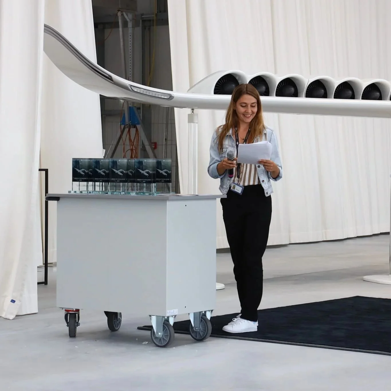 Woman holding a microphone and reading from papers standing near a small wheeled table with trophies on top, in front of a large aircraft engine model.