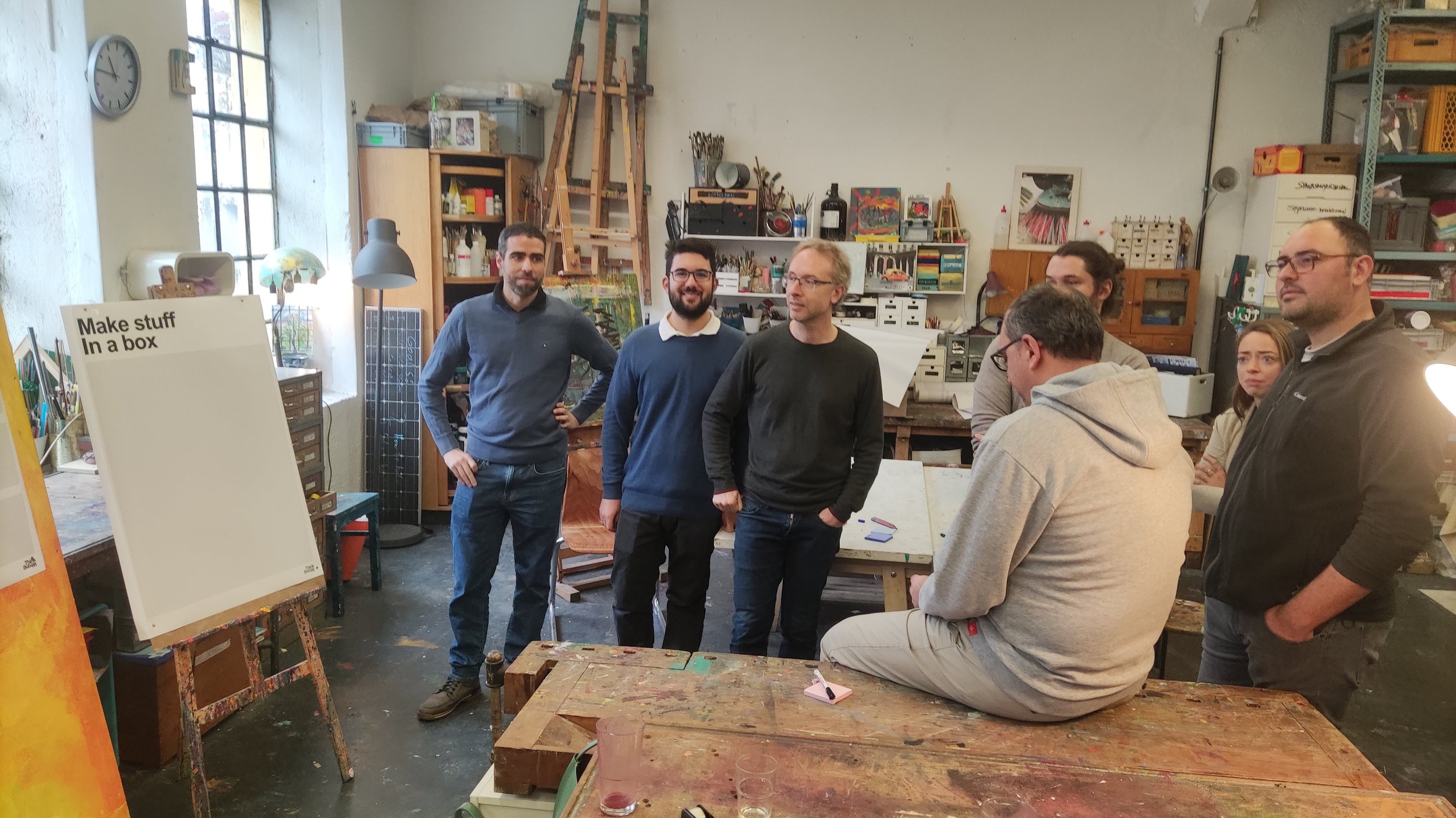 Group of people in an art studio or workshop with paintings, and art supplies visible.