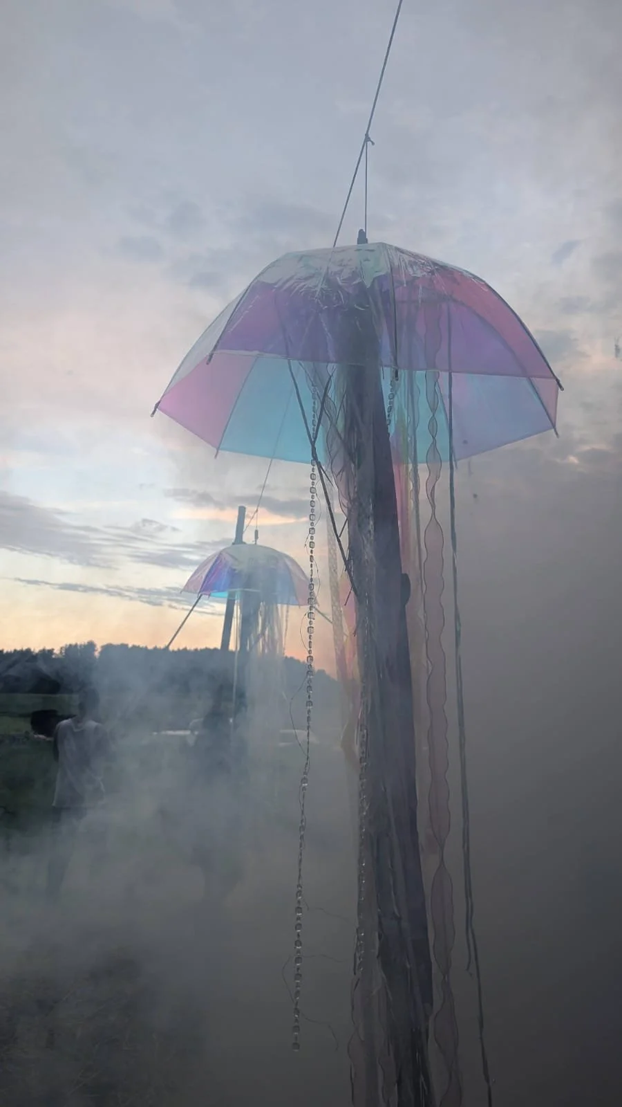 Colorful, transparent umbrellas hanging from a pole against a cloudy sky during dusk, with smoke and a person in the background.