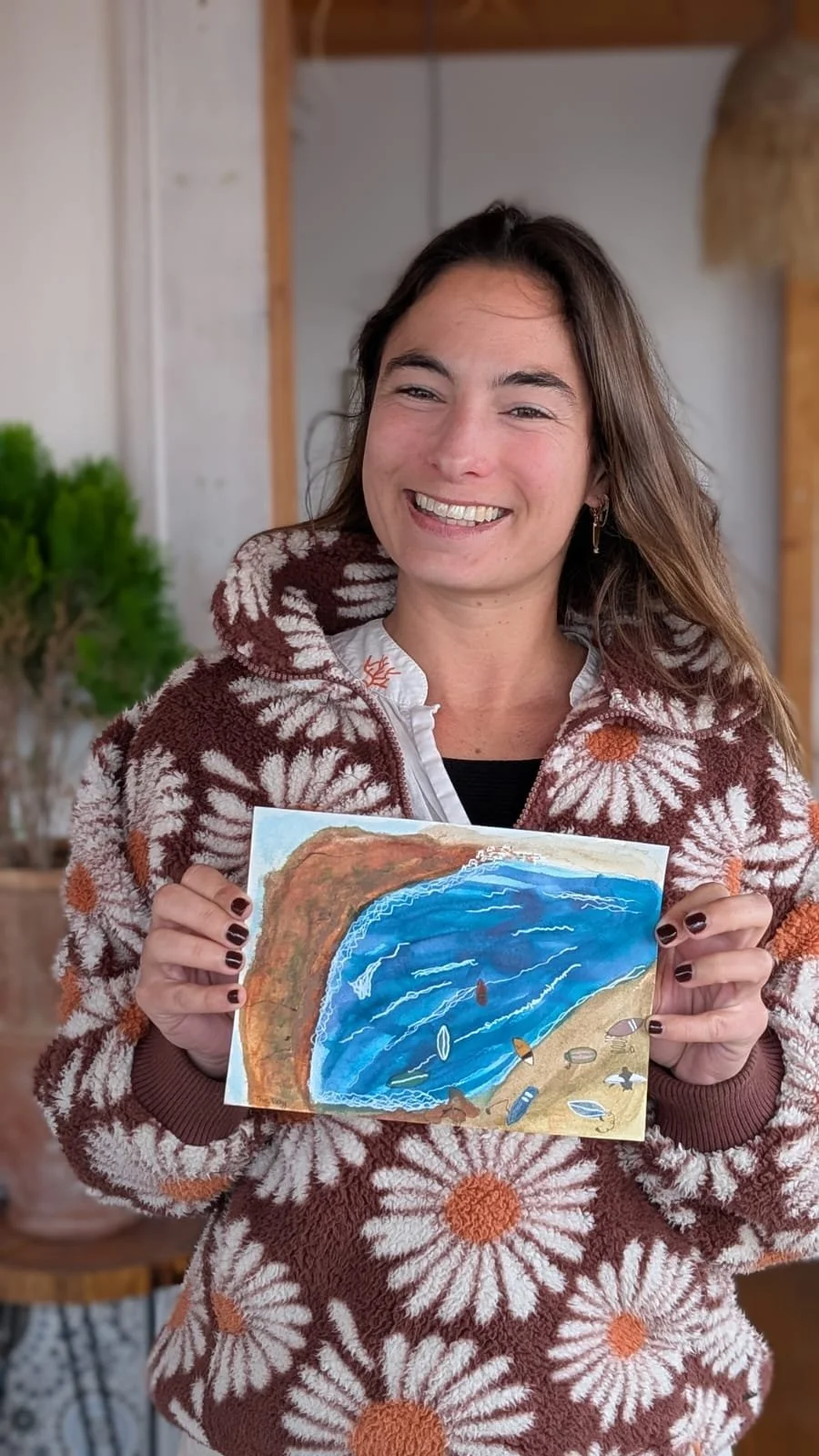 A woman smiling and holding a painting of a coastal scene with a cliff, ocean, and sky, wearing a patterned fleece jacket with sunflowers.