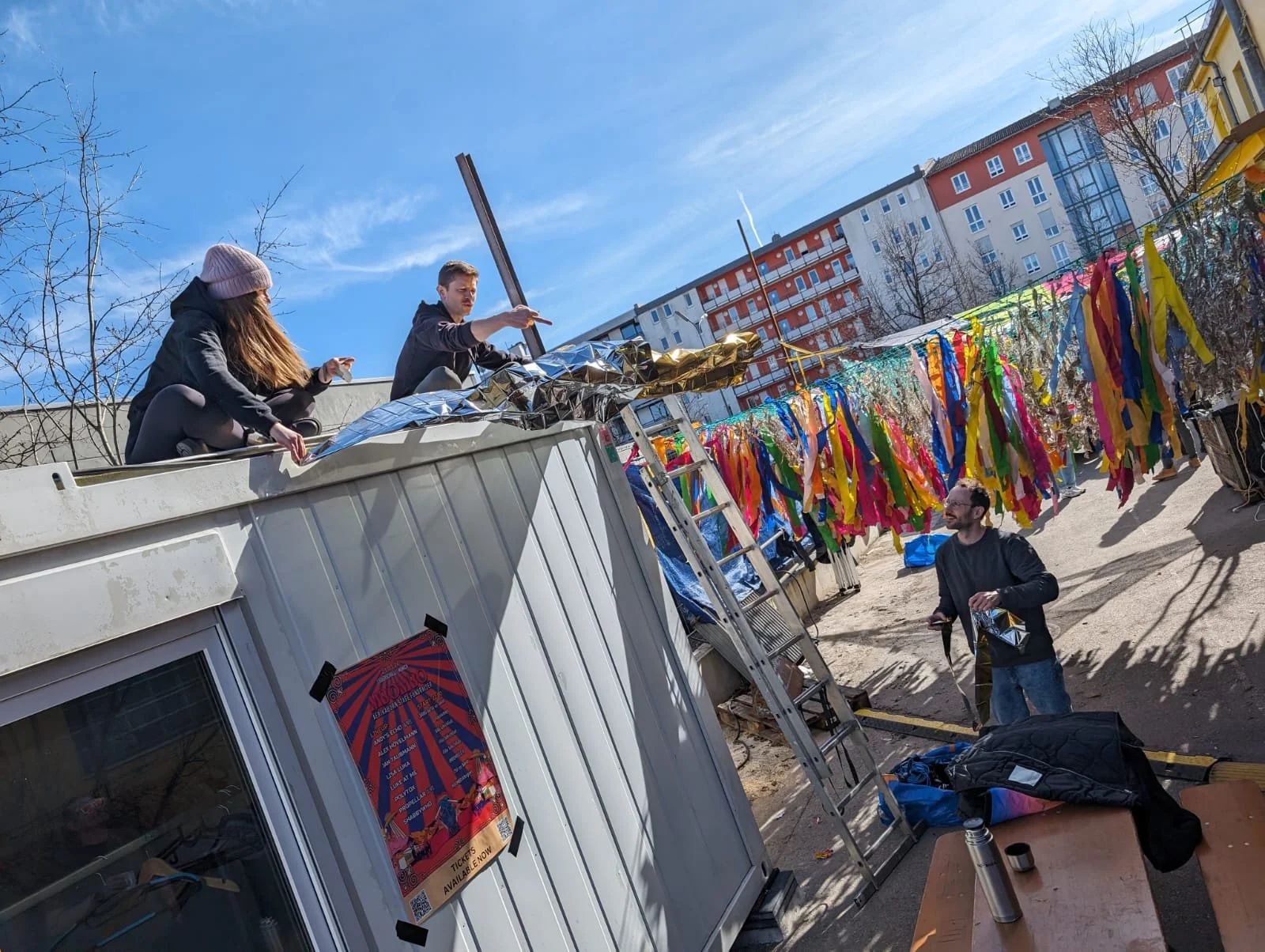 People setting up colorful decorations on a sunny outdoor rooftop with a ladder. Two women are on the roof and a man stands below with a phone.