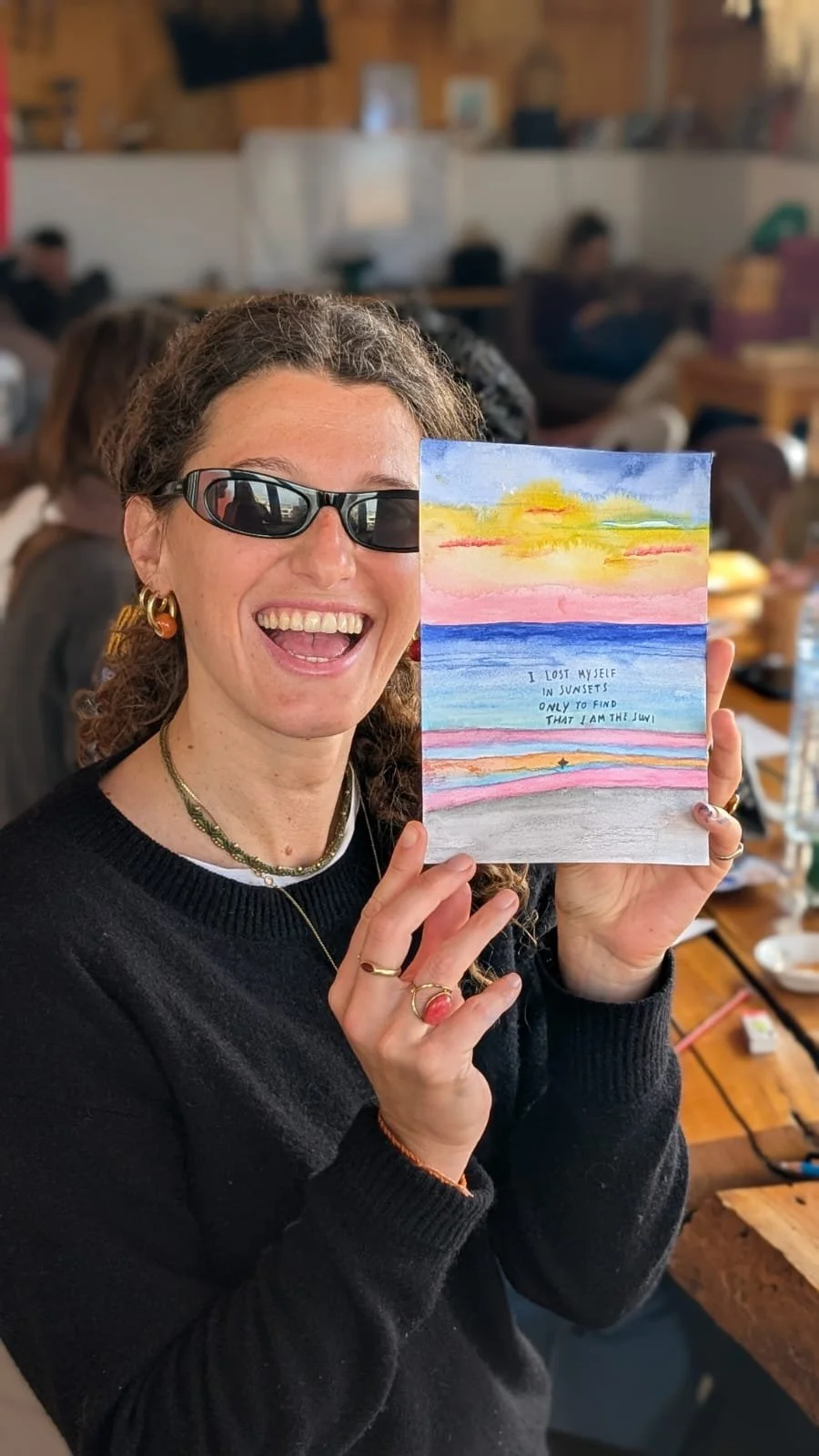 Woman holding colorful sunset painting with a quote about sunsets, smiling in a cozy indoor setting with other people in the background.