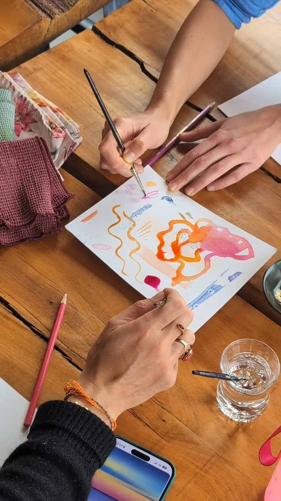 Two people creating an abstract watercolor painting with pink, orange, and purple colors on paper, on a wooden table with art supplies and a glass of water.
