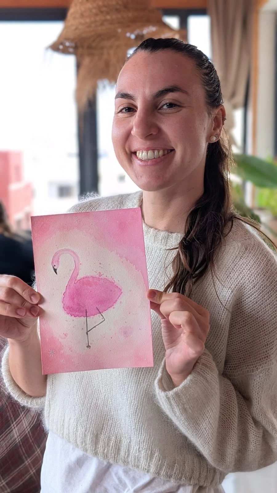 A smiling woman holding a pink card featuring a flamingo watercolor painting, sitting indoors near a window with natural light.
