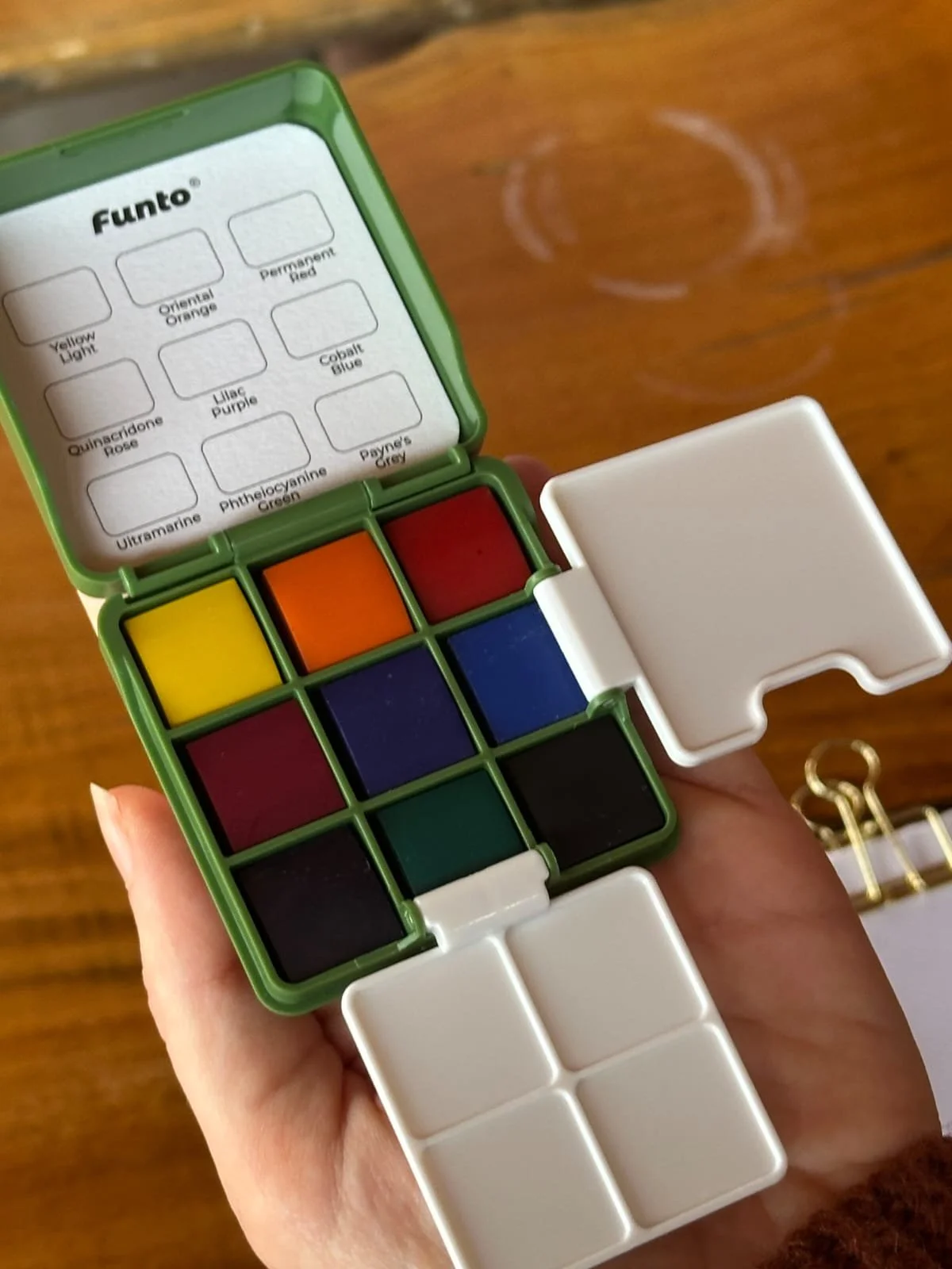 Open watercolor palette with 12 colors including yellow, orange, red, blue, green, and black, with a white mixing area.