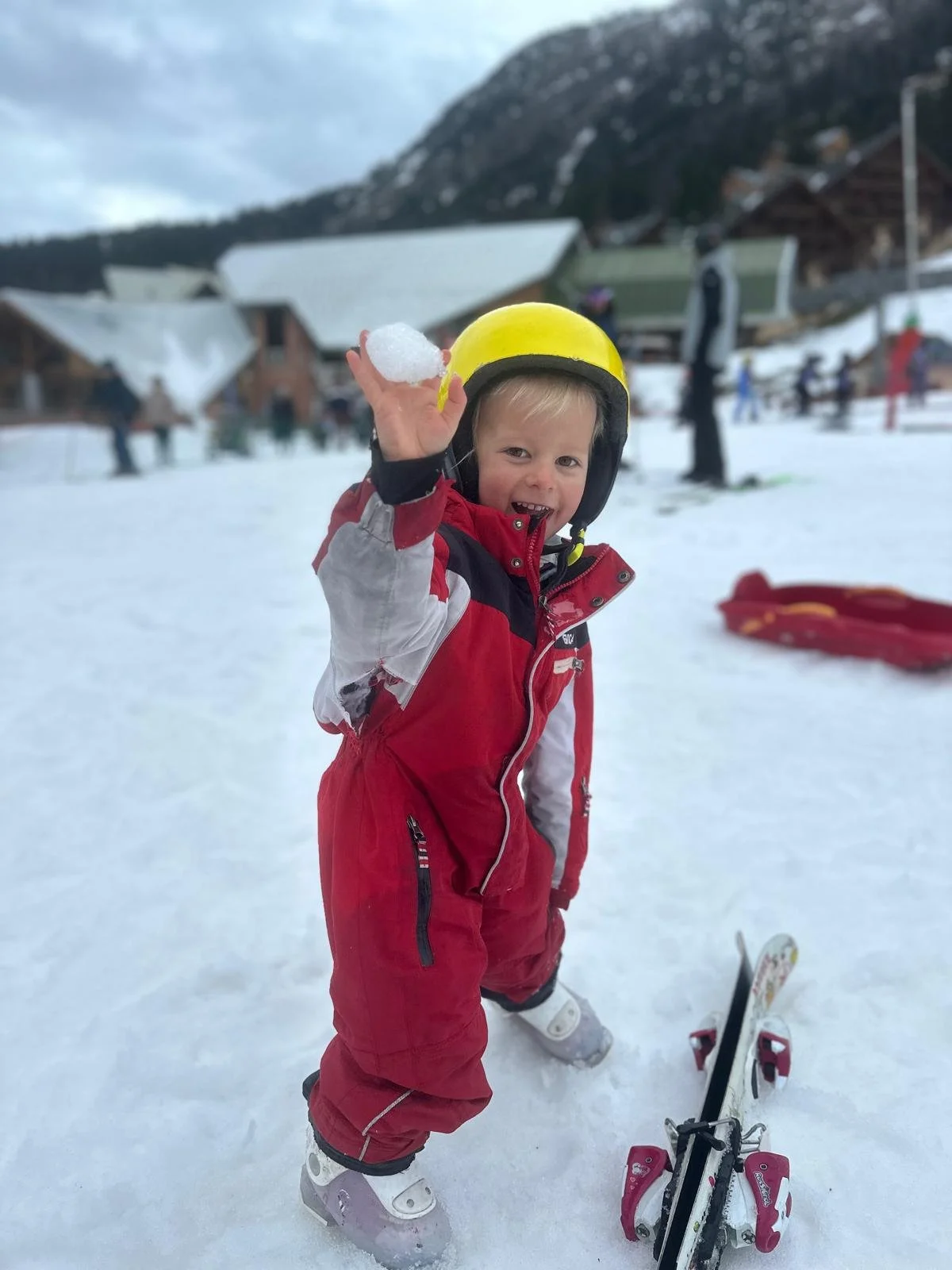 Young boy in red and white snow gear with a yellow helmet holding snow above his head on a snowy mountain with other skiers and slopes in the background.