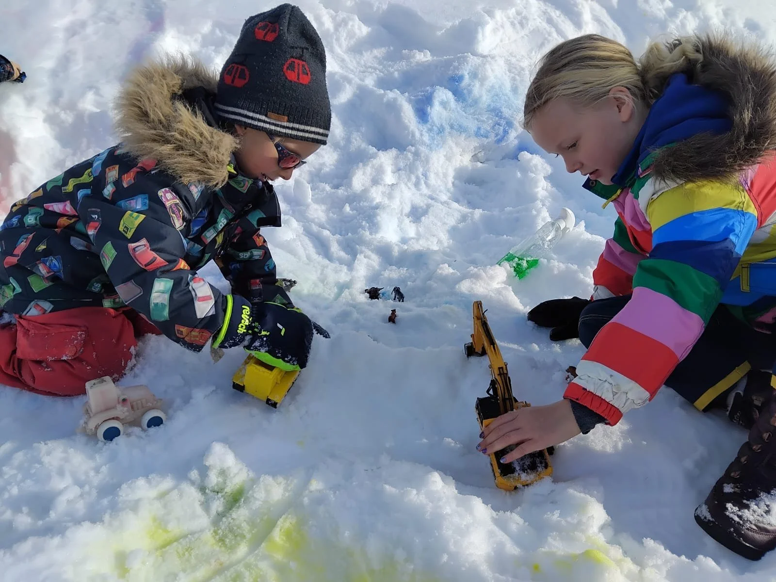 Two children dressed in colorful winter jackets playing in the snow with toy construction vehicles, surrounded by snow-covered ground.