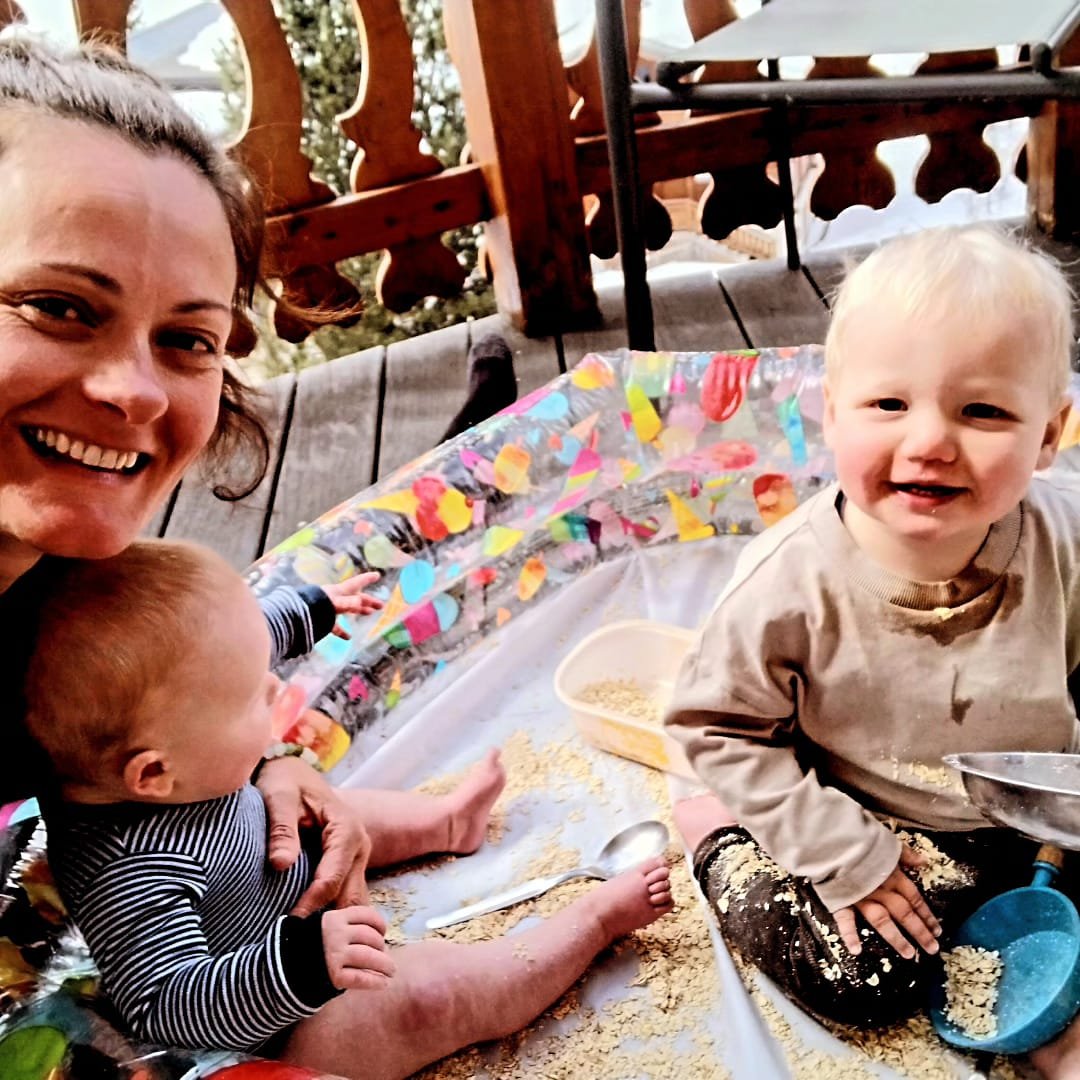A smiling woman with two young children on a colorful outdoor patio. The children are playing with oatmeal or similar food, with one child sitting on the blanket and the other being held. The scene is cheerful and messy, with breakfast or snack time activity.