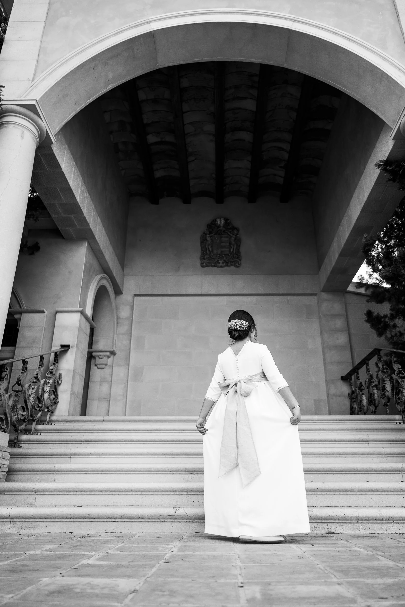 Mujer vestida de blanco con un moño en el cabello, vista de espaldas, subiendo unas escaleras frente a un edificio con arcos y escudo en la pared.