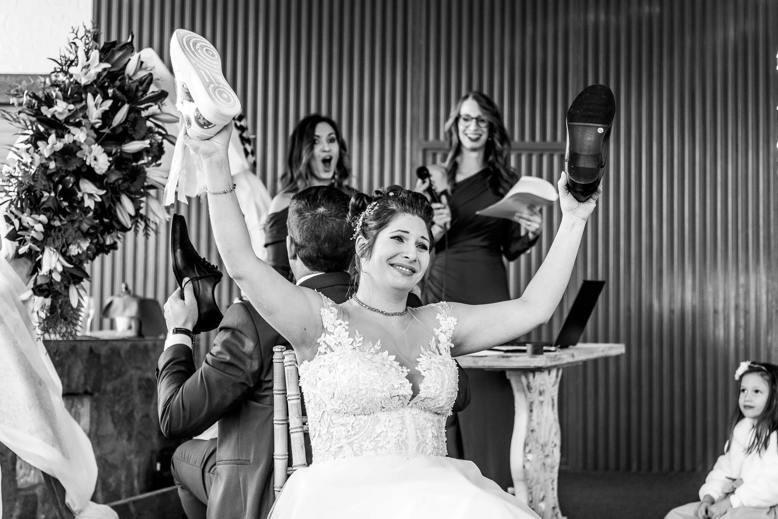 Mujer con vestido de novia levantando los brazos en una celebración de boda, rodeada de amigos y familiares en un ambiente interior con paredes de madera.