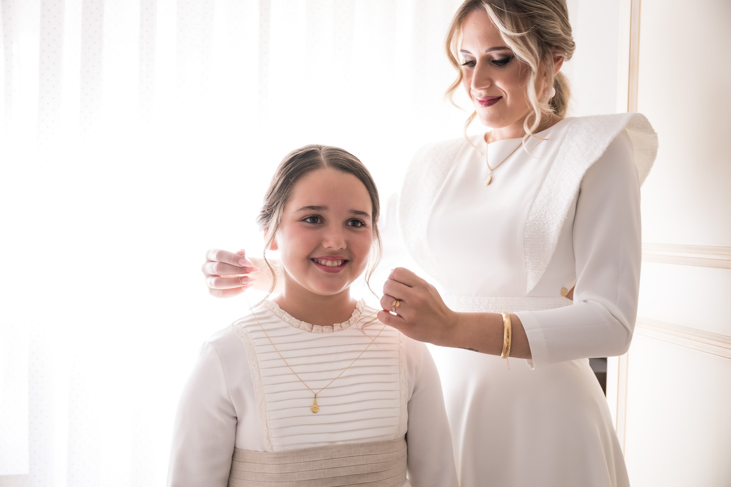 Una mujer mayor en un vestido blanco colocando un collar en un niño de cabello oscuro y vestido a rayas blancas y rosas, frente a una ventana con cortinas blancas.