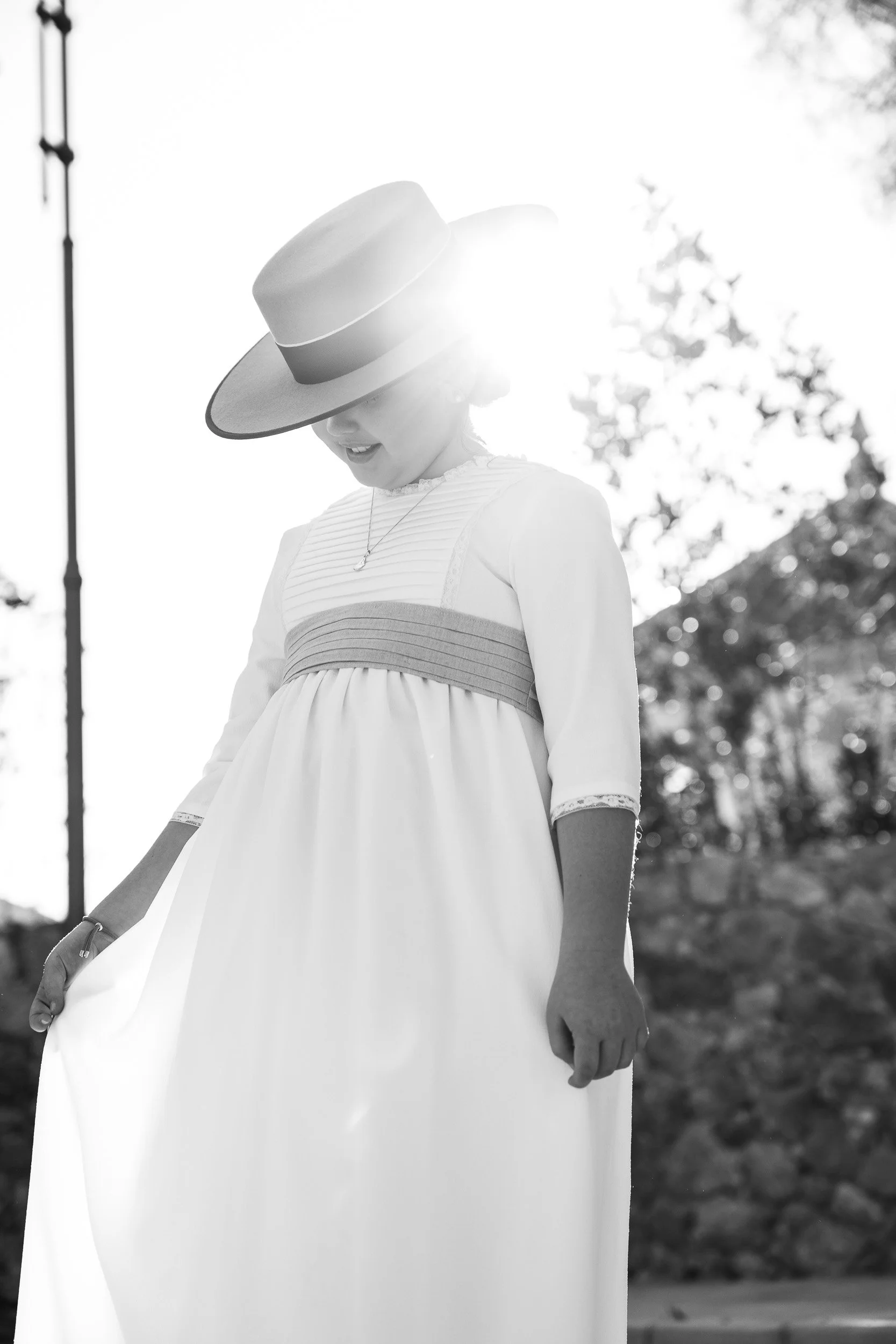 Mujer joven usando un sombrero grande y vestido largo, sonriendo al exterior en un día soleado con árboles en el fondo.