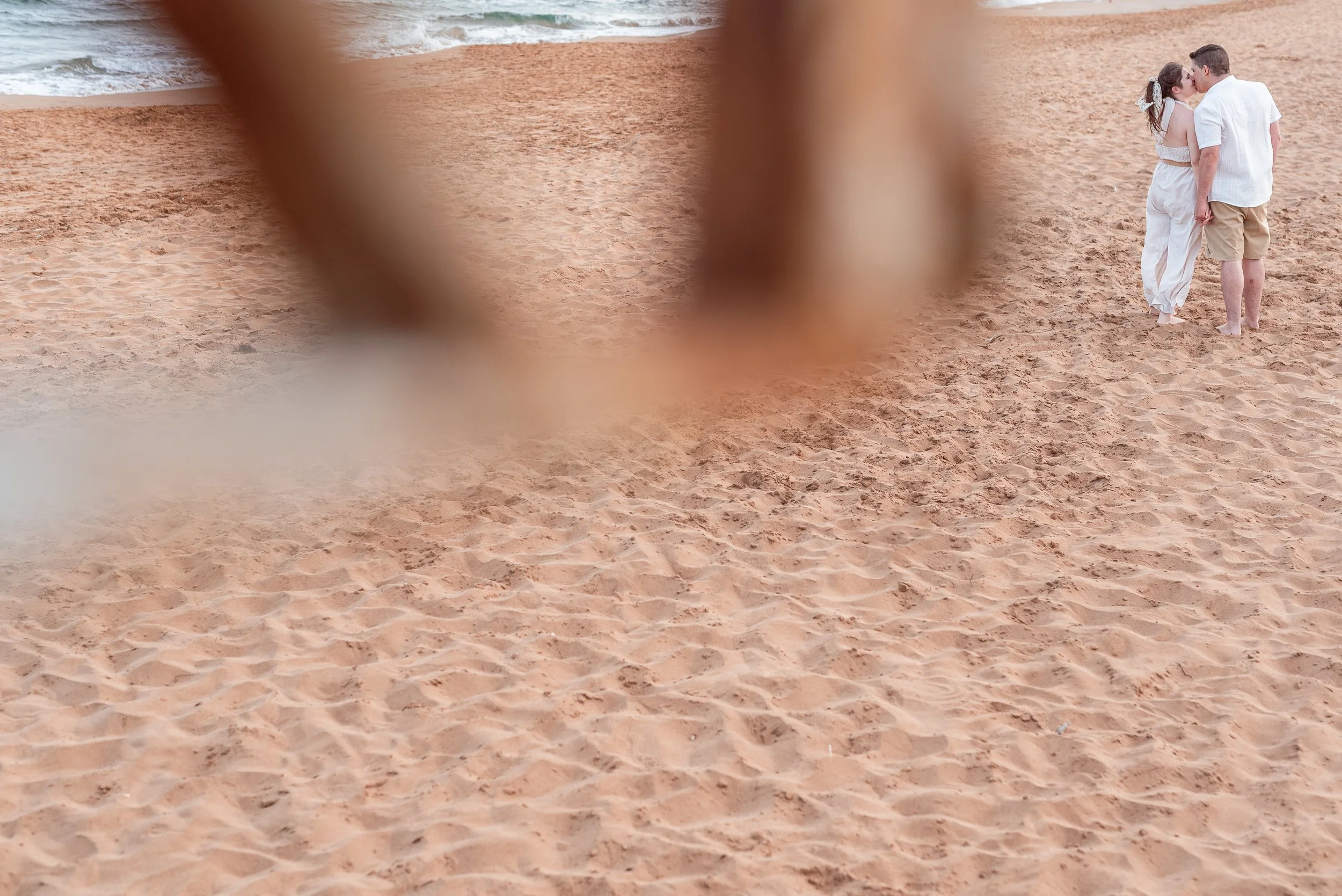 Pareja besándose en la playa, vista parcialmente bloqueada por una estructura de madera.