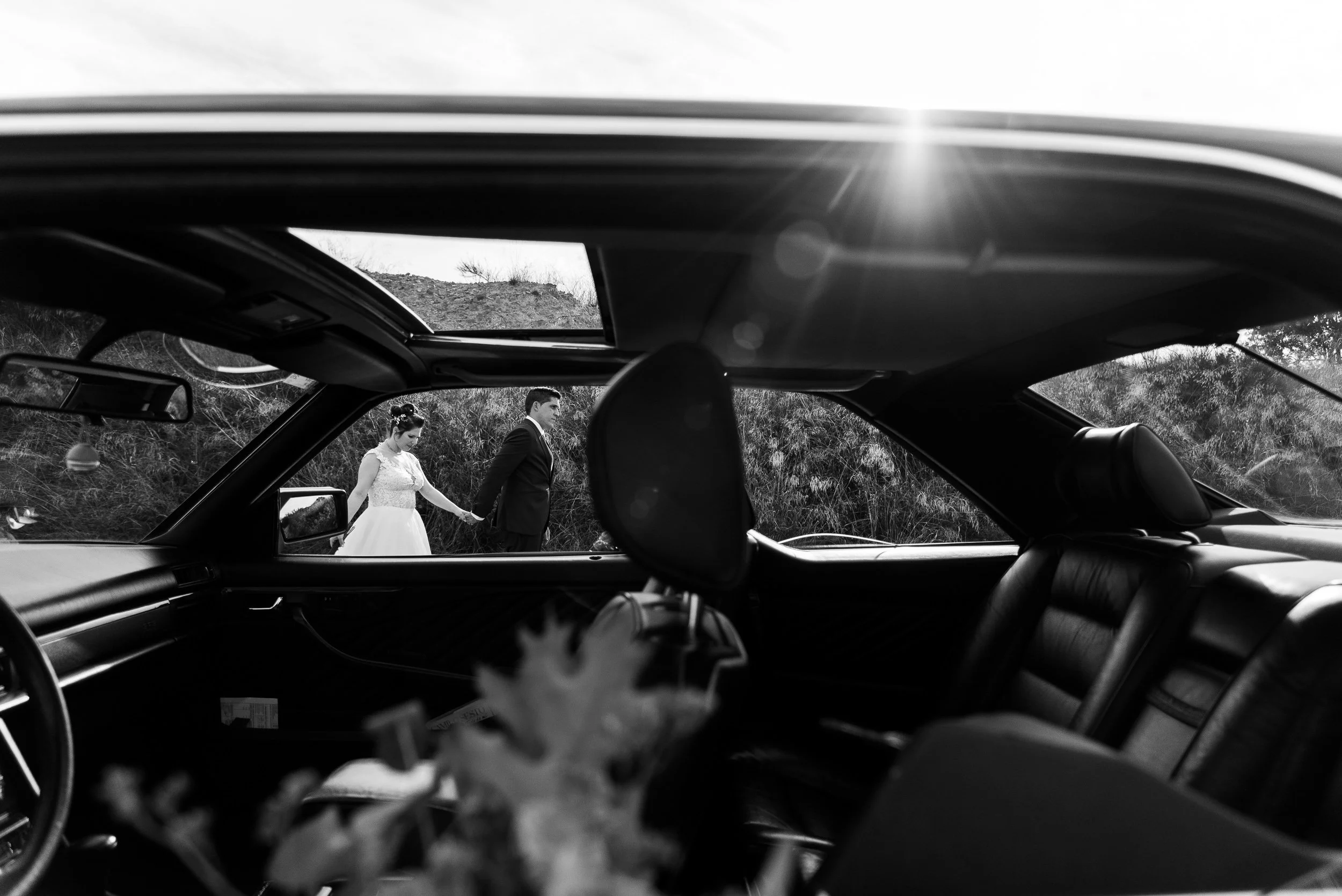 Una pareja vestida de boda caminando por un sendero natural, vista desde el interior de un auto con el techo abierto en un entorno exterior natural.