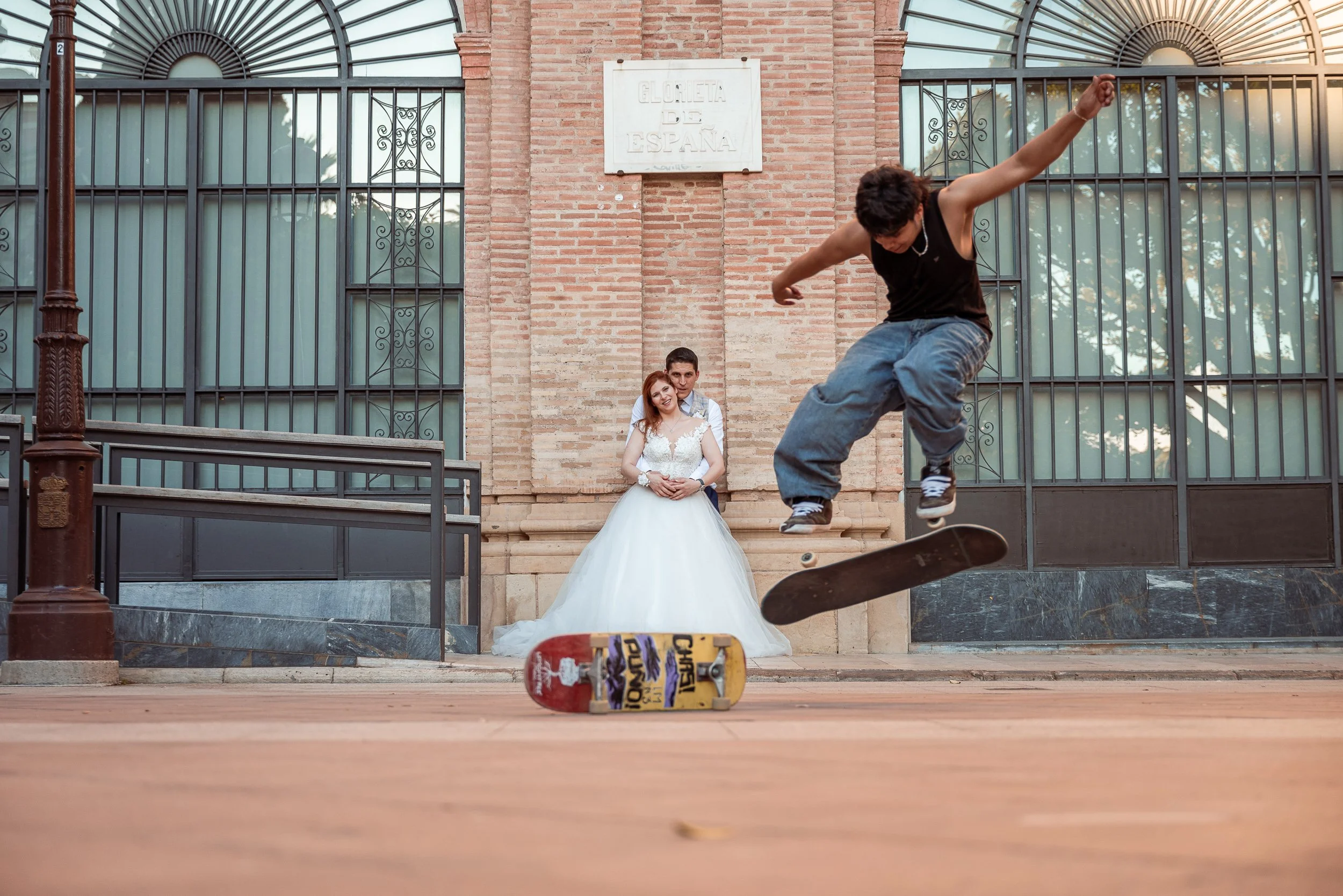 Un joven haciendo trucos en skate frente a una pareja con vestimenta de boda en un espacio público con puertas de rejas y paredes de ladrillo.