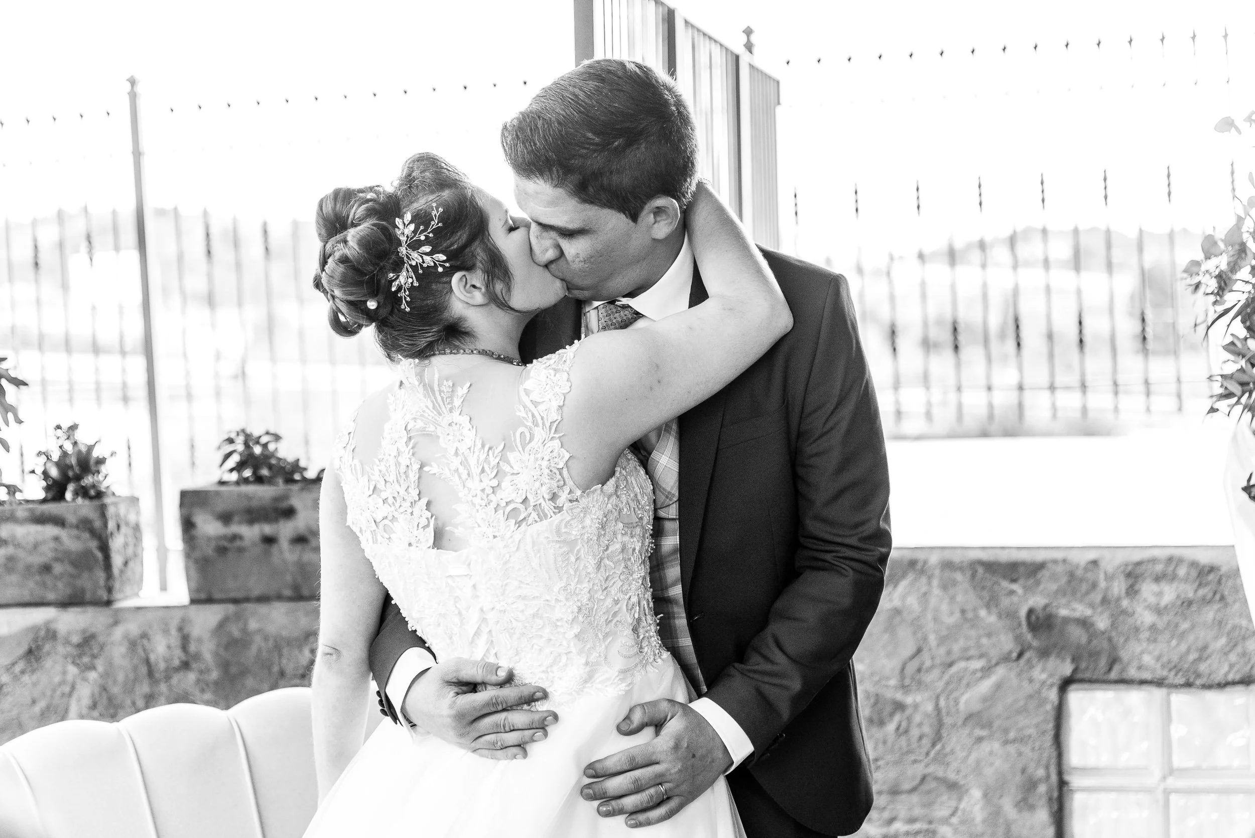 Una pareja recién casada se besa con los ojos cerrados en un entorno exterior, ella lleva un vestido de novia y él un traje. La escena capta un momento romántico y feliz en su boda.