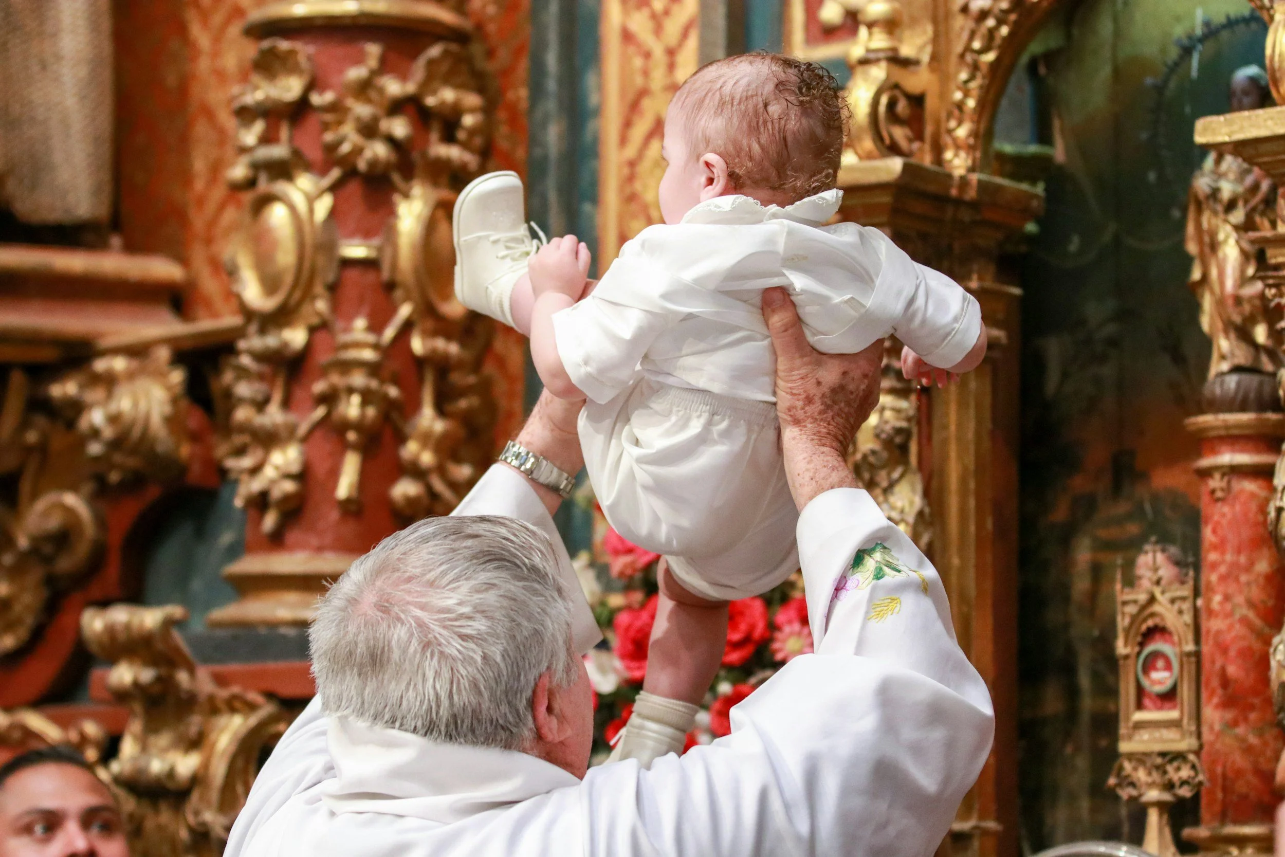 Un niño pequeño de bautizo está siendo levantado en el aire por un cura hacia el altar en una iglesia.