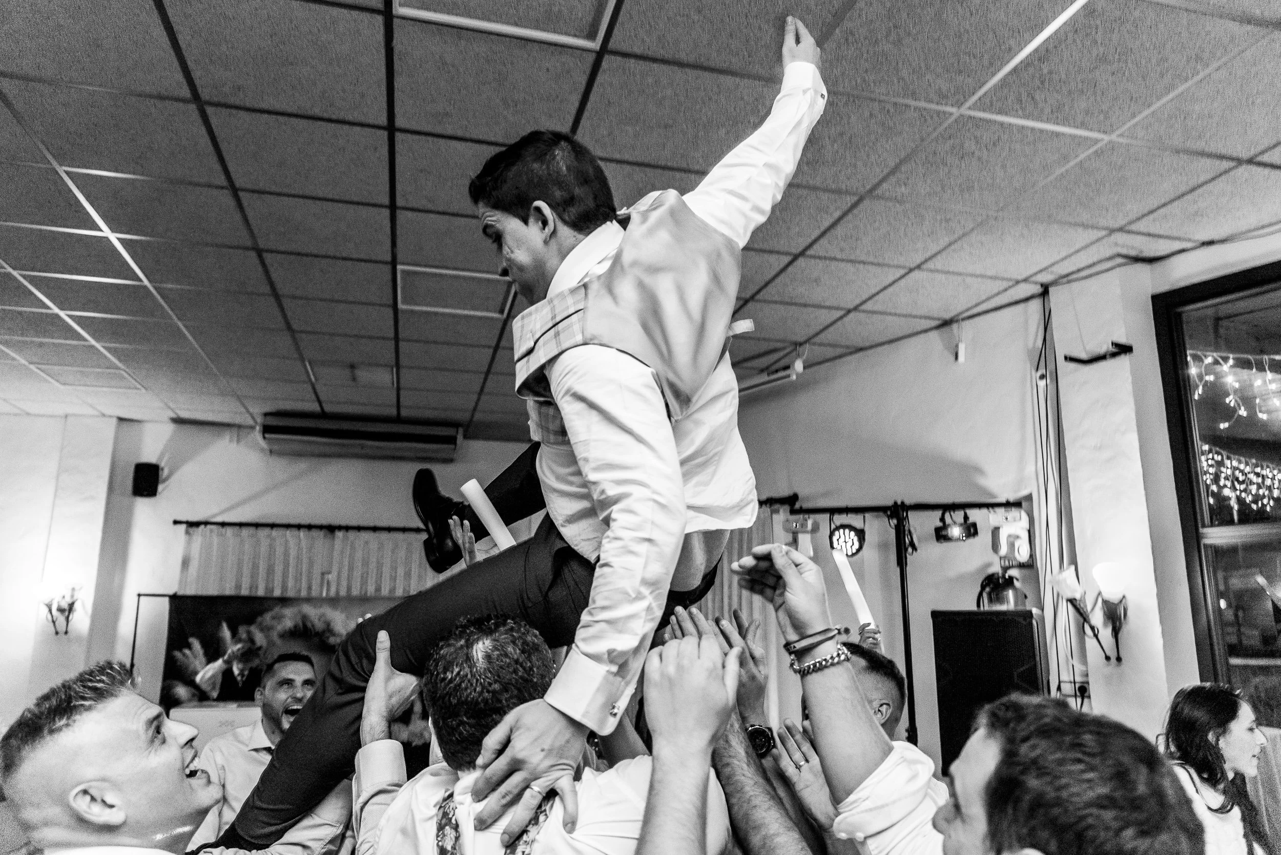 Hombres celebrando en una reunión, levantando a un compañero que baila en medio del grupo, en un ambiente interior.