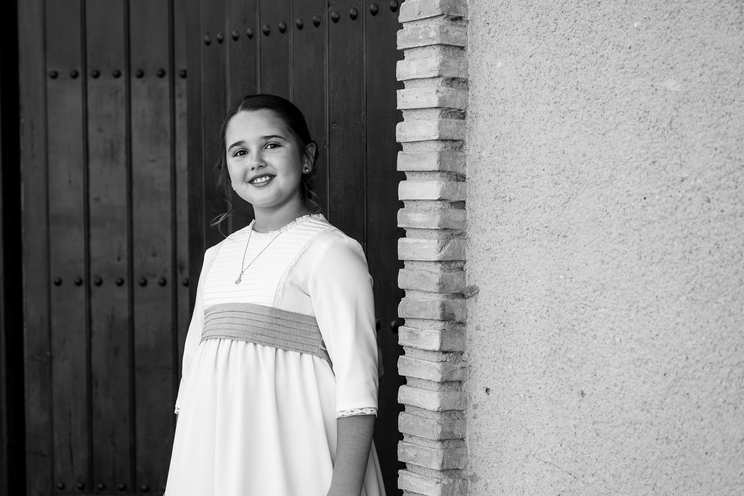 Niña sonriendo, con cabello oscuro y liso, vestida con un vestido blanco con detalles en el cuello, de pie junto a una pared de ladrillos y una puerta de madera oscura.