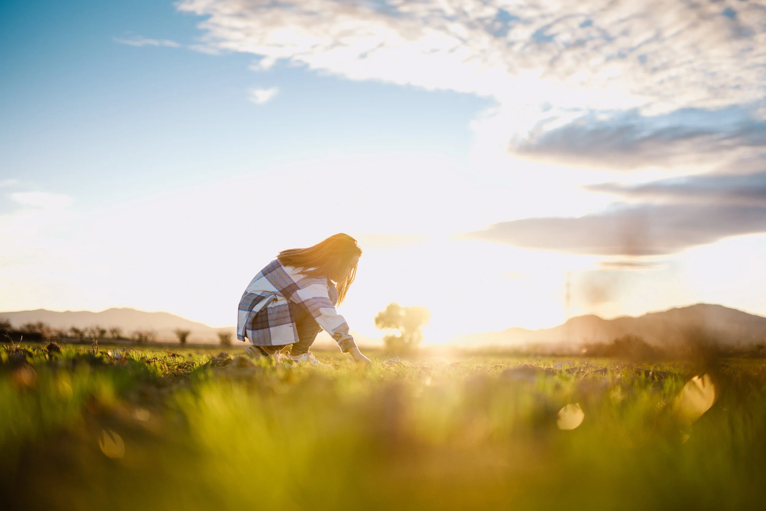 Una niña al aire libre en un campo, arrodillada y cuidando las plantas al atardecer, con montañas y cielo con nubes en el fondo.