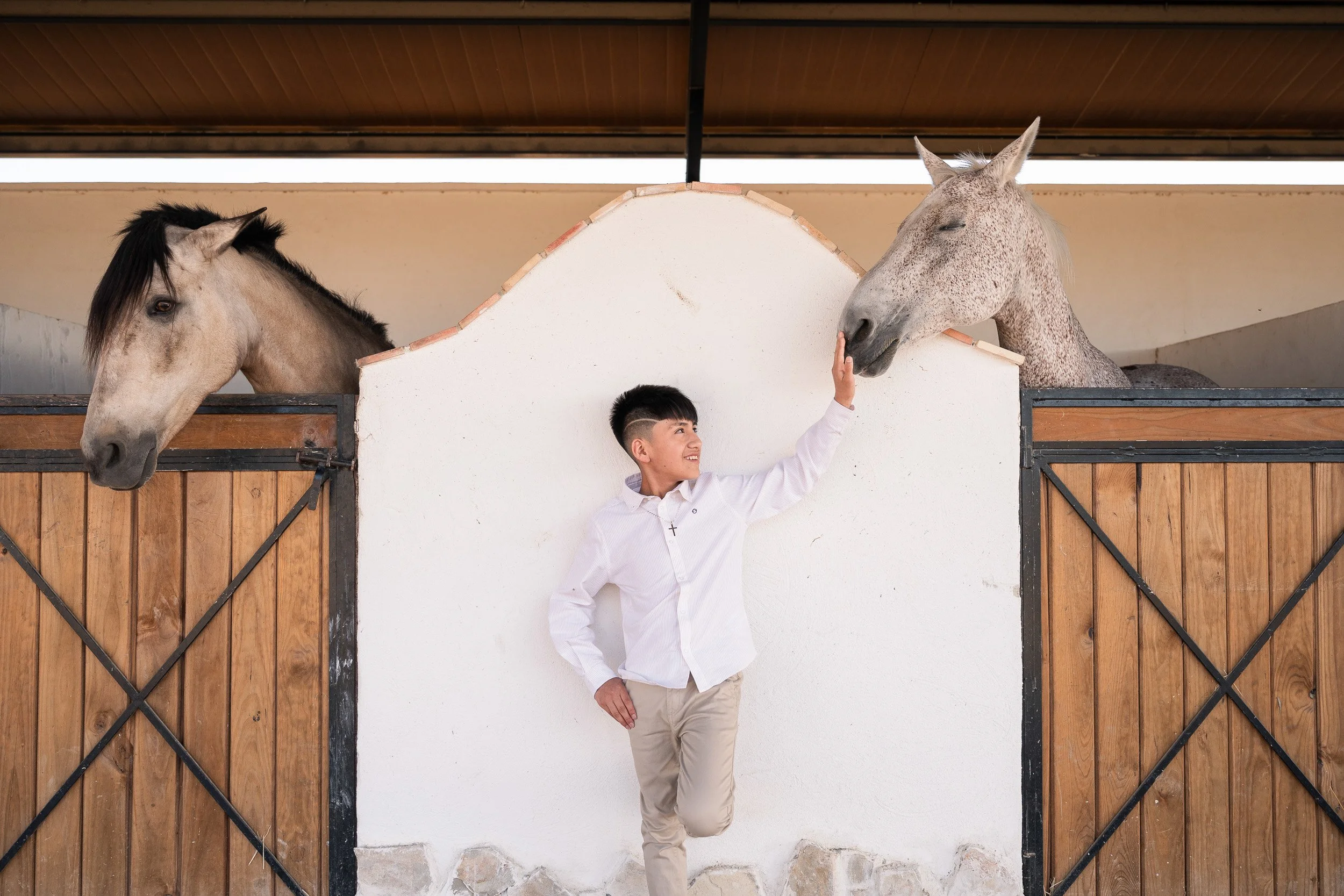 Un niño con camisa blanca y pantalones beige acaricia la cara de un caballo gris con manchas negras, en un establo de madera con un muro blanco y techo de madera.