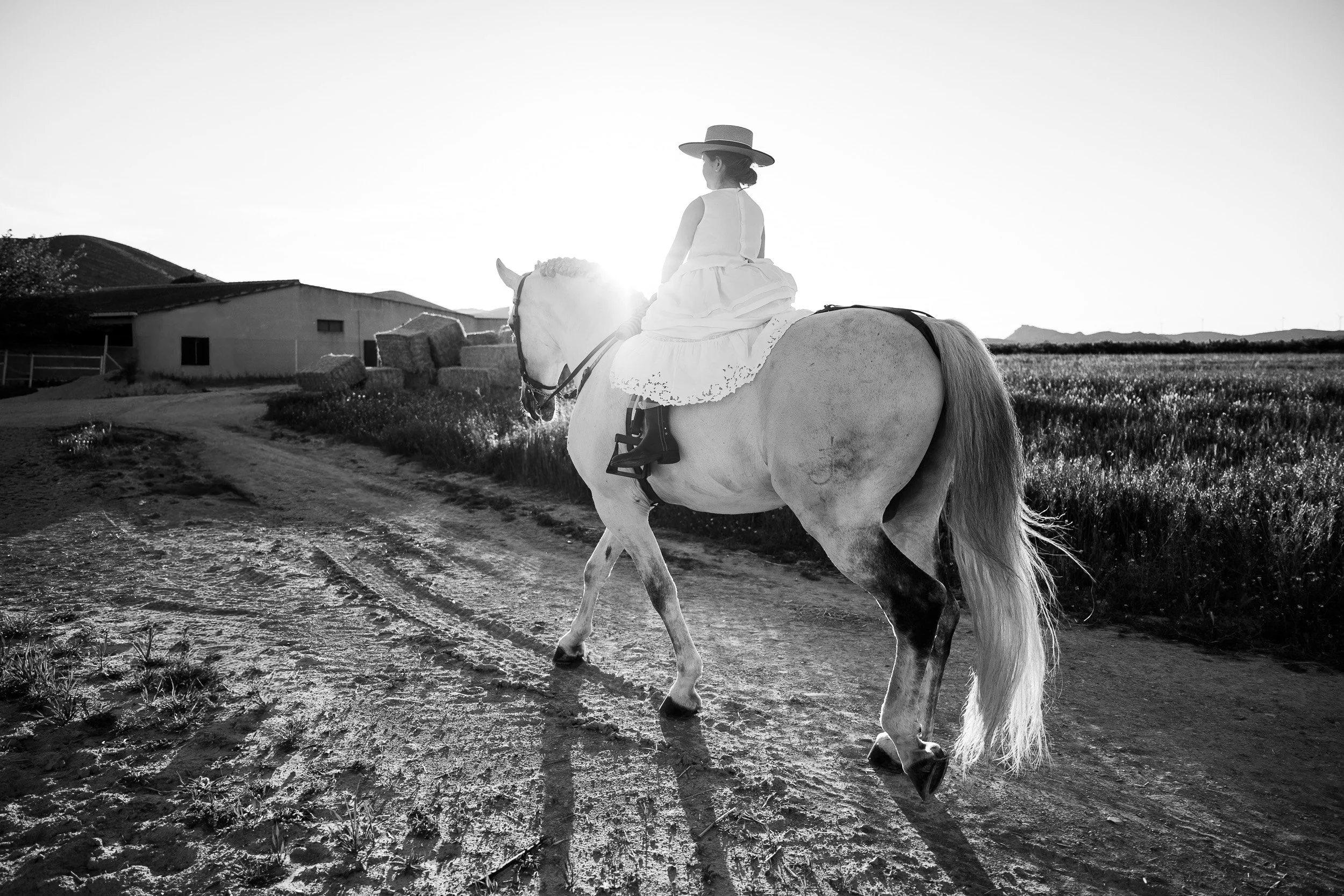 Una mujer montando un caballo en un campo, en un entorno rural, con edificios y colinas al fondo y el sol en el horizonte, en blanco y negro.