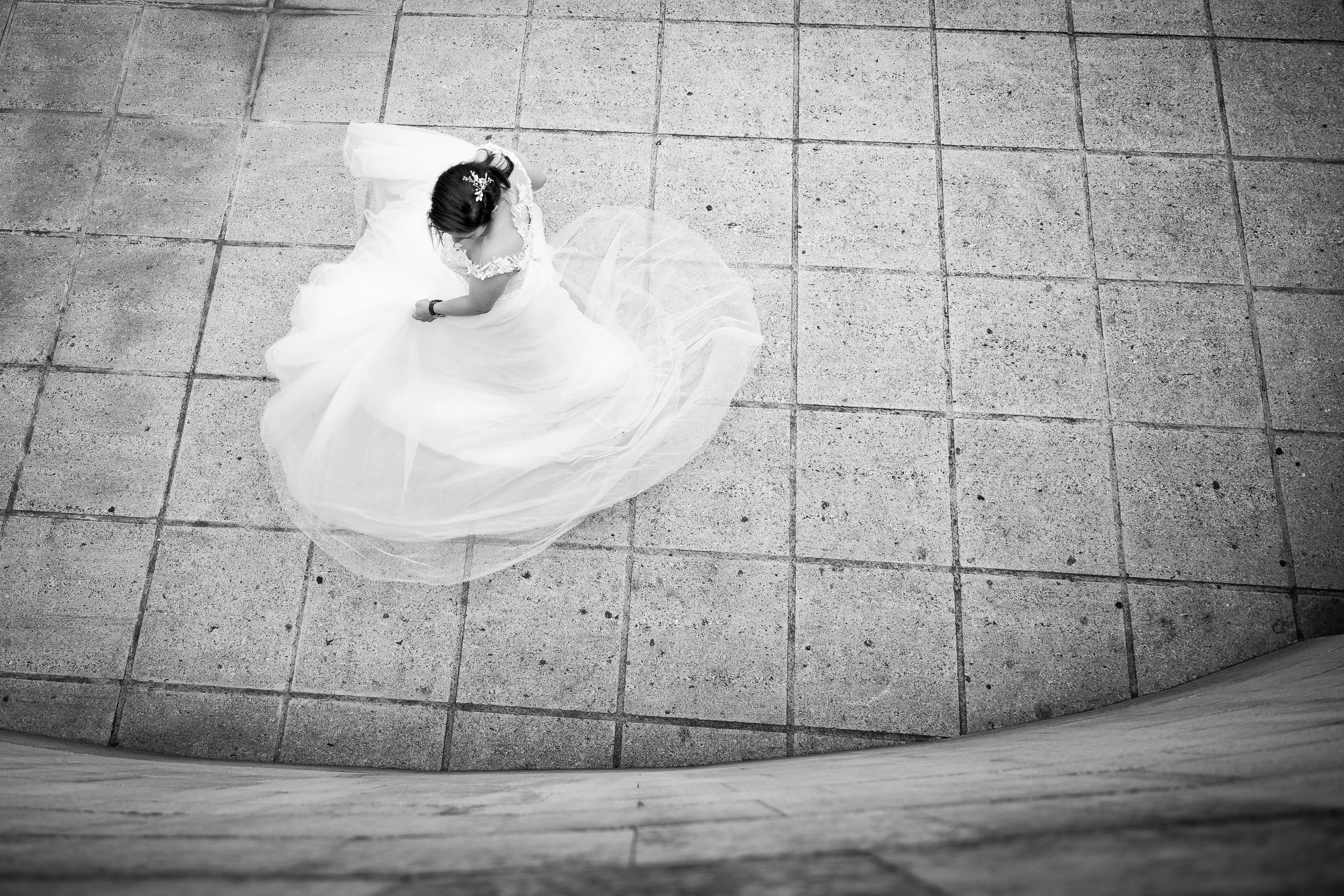 Mujer vestida con vestido de novia y velo con una diadema, vista desde arriba, en un piso de baldosas de cemento.