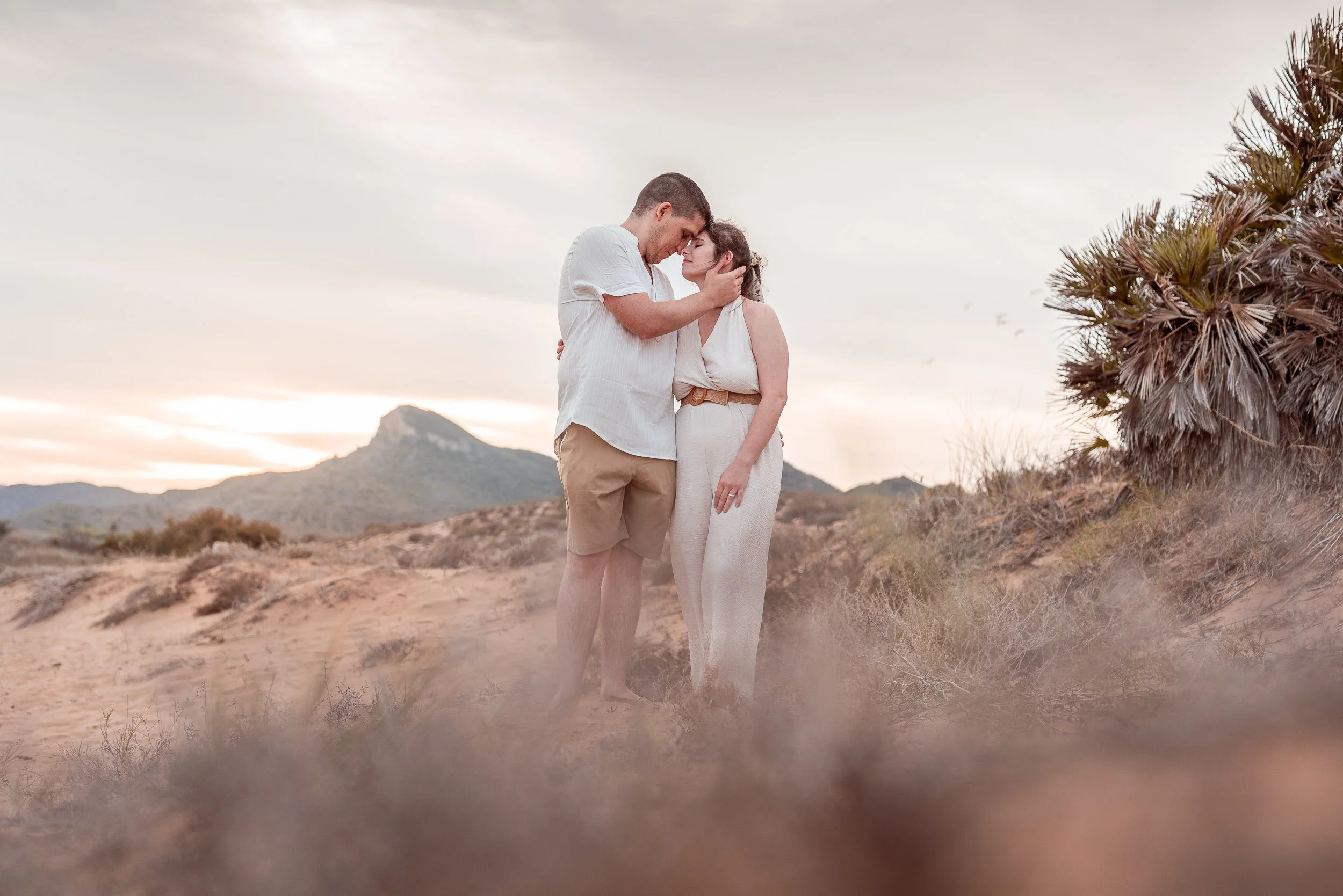 Una pareja se abraza y se besa en un paisaje desértico durante el atardecer, con montañas y cactus en el fondo.