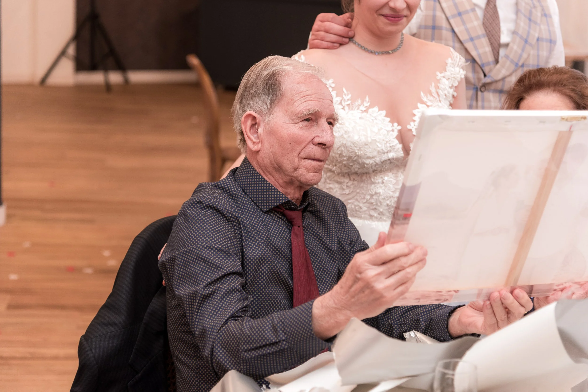 Un anciano hombre con camisa negra de puntos rojos y corbata roja sentado en una mesa, leyendo un libro, en un entorno de celebración con otros adultos en el fondo.