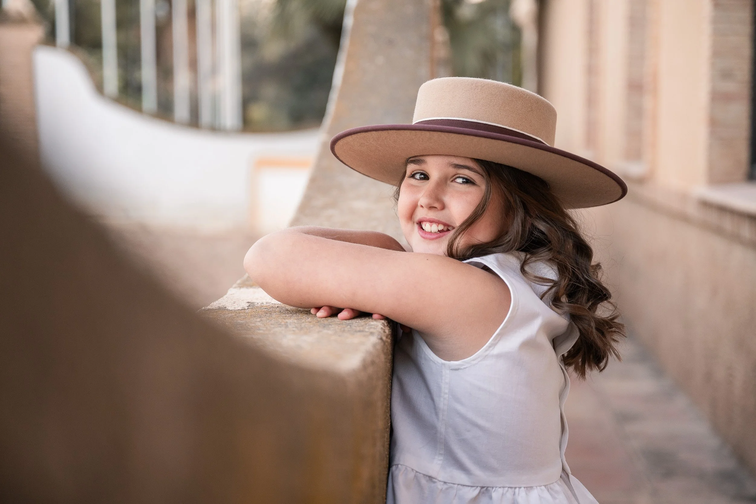 Niña con sombrero grande de ala ancha recostada en un muro de piedra, sonriendo y mirando hacia la cámara, con fondo de arquitectura y vegetación.