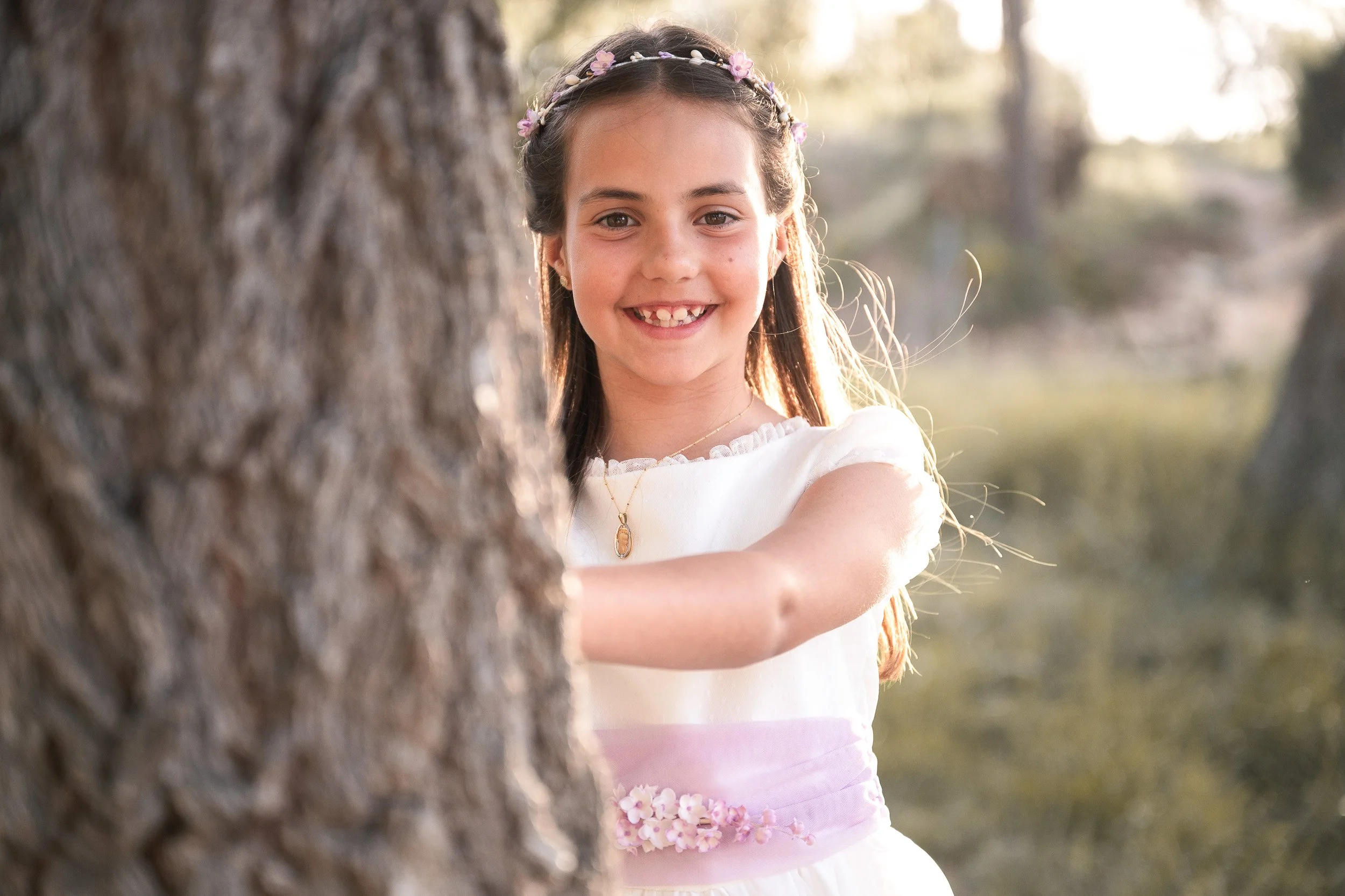 Una niña sonriente vestida de comunión, apoyada en un árbol, en un entorno natural con luz suave del atardecer.