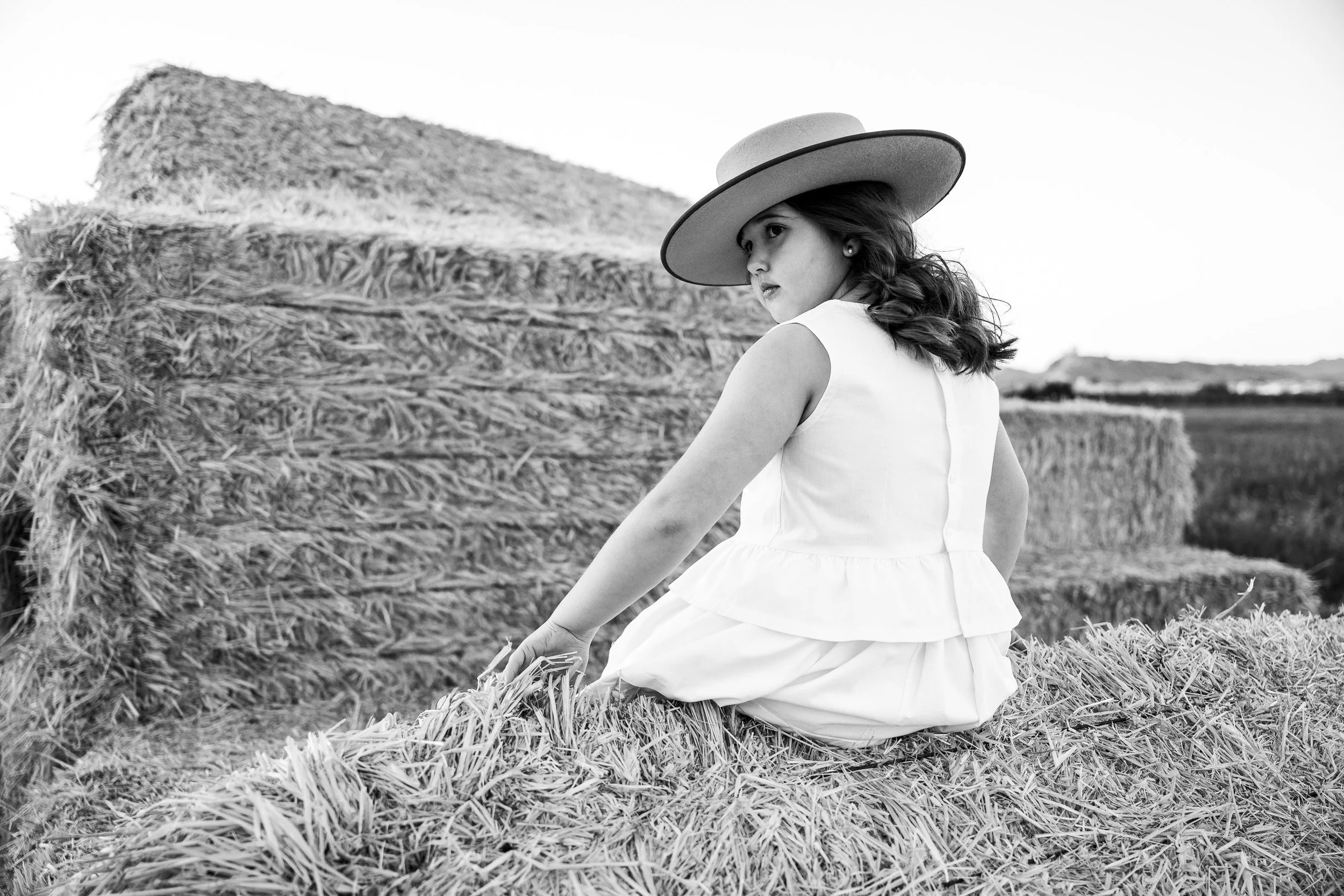 Joven mujer en vestido blanco y sombrero grande sentada sobre paja en campo agrícola, mirando hacia atrás en fotografía en blanco y negro.