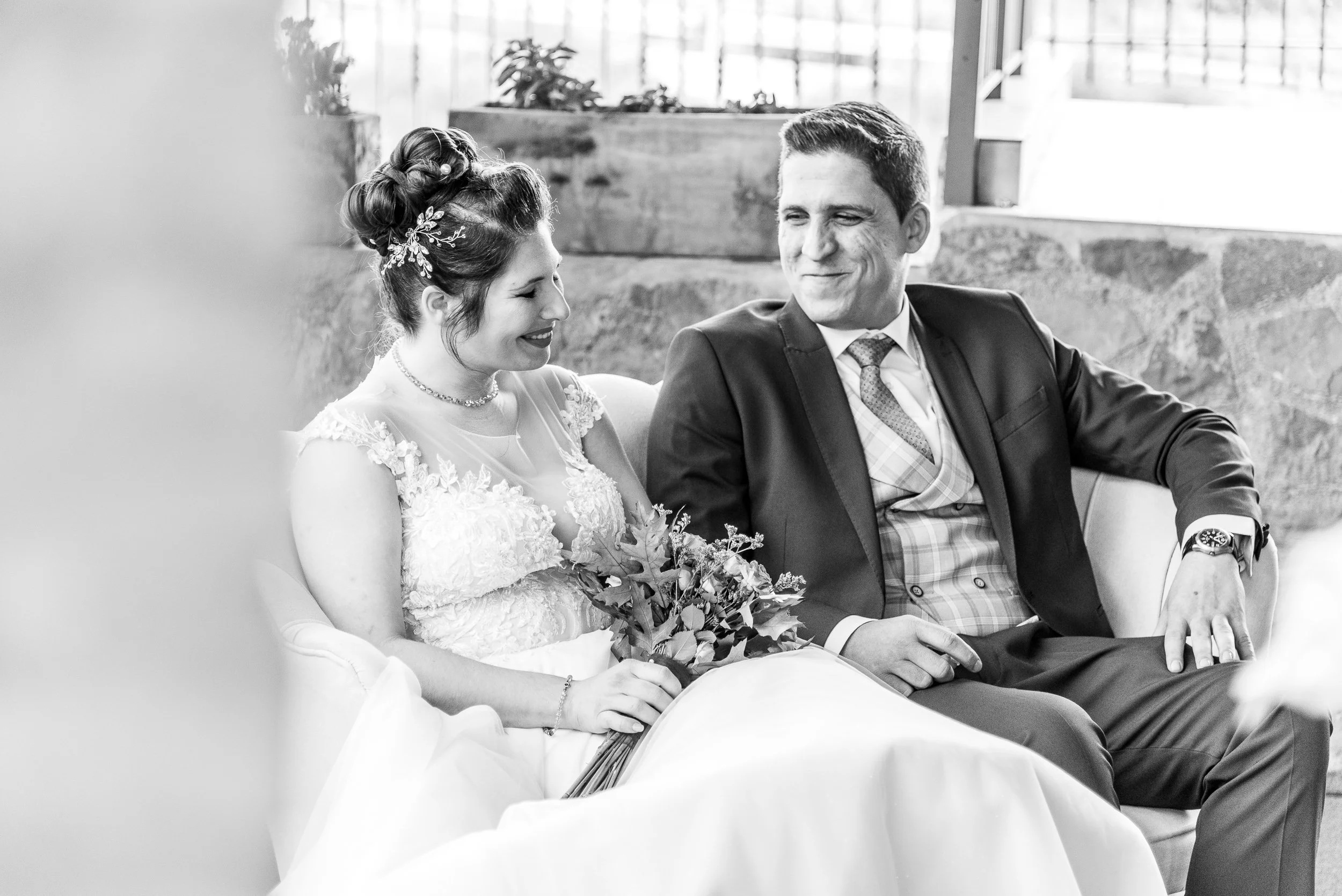 Pareja de novios en boda, la novia con vestido y tocado, el novio con traje, sentados en un banco, sonriendo y sosteniendo un ramo de flores, en un entorno interior con ventanas y paredes de piedra.