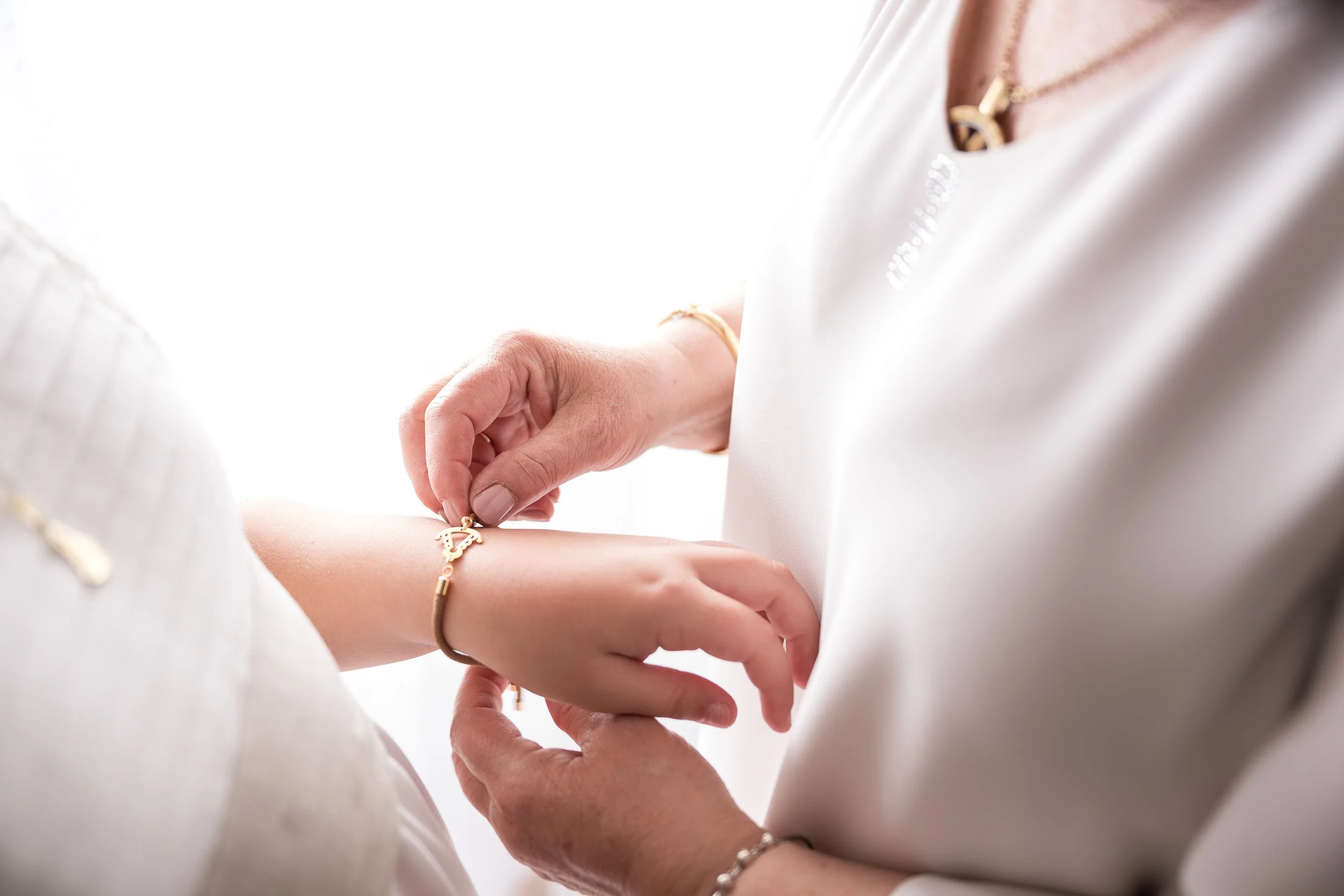 Persona colocando una pulsera en la muñeca de otra persona, ambas usando ropa blanca y accesorios dorados.