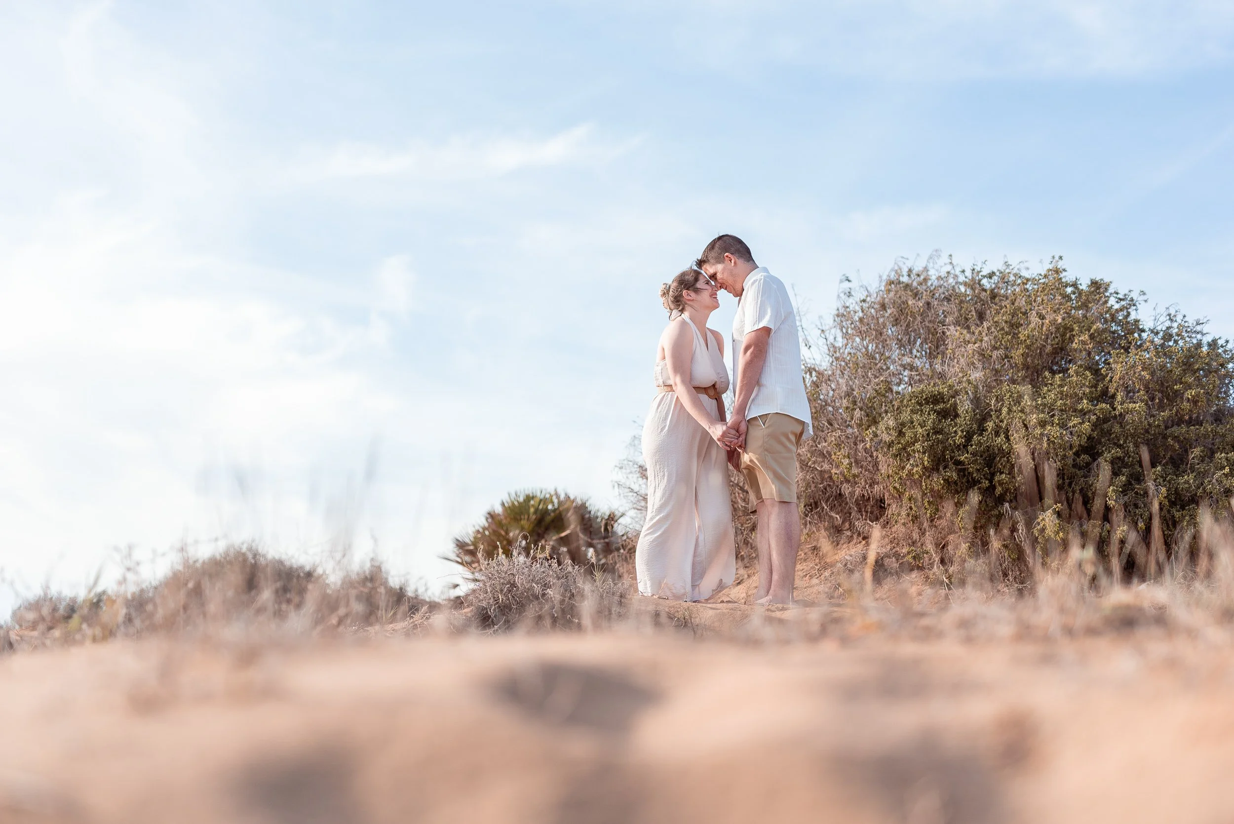 Una pareja se encuentra en un campo seco, de fondo hay árboles y un cielo azul con algunas nubes, parecen felices y están muy cercanos, tocándose con las frentes y entrelazando las manos.
